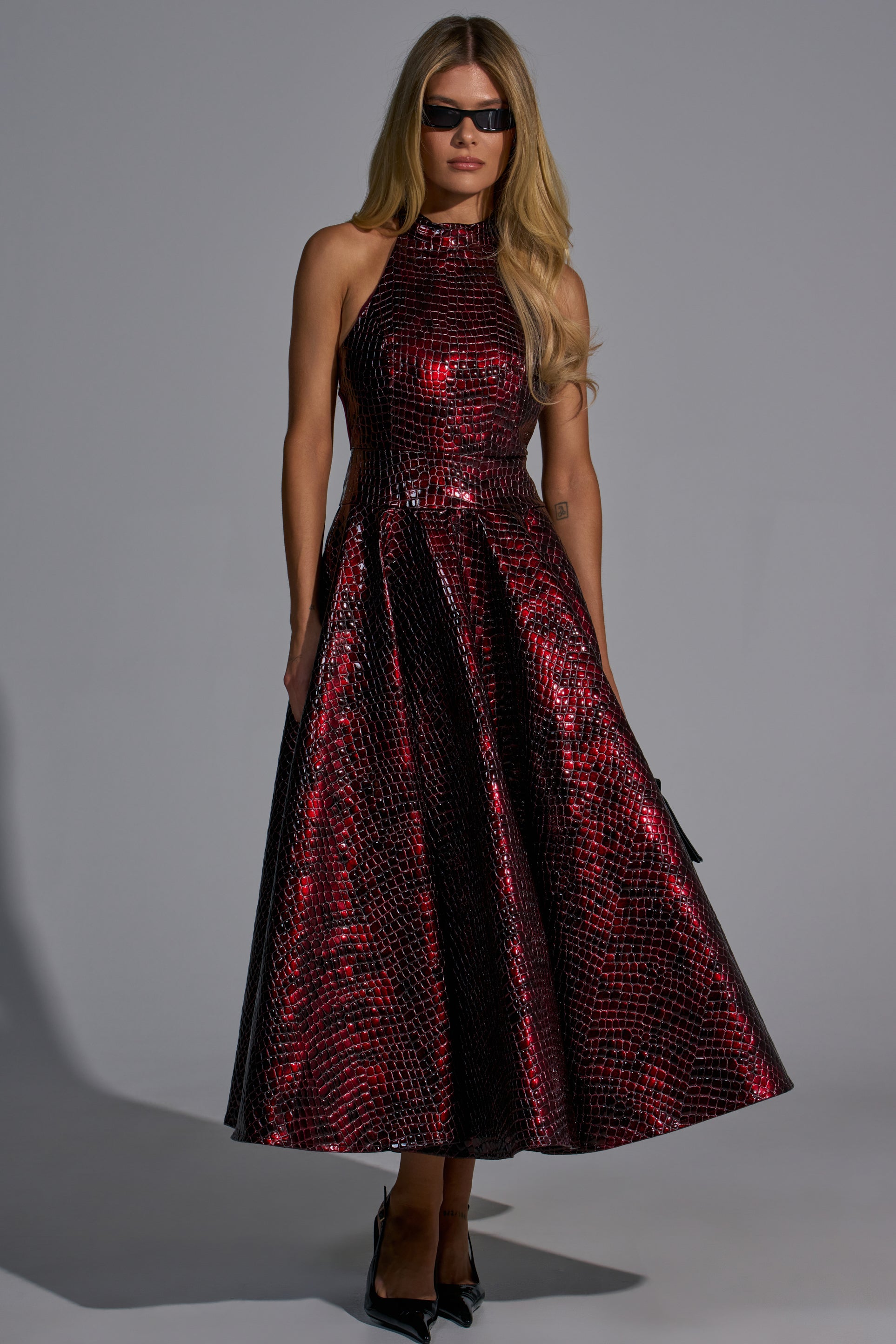 A woman wearing a VEE FAUX LEATHER CROC MIDI DRESS IN BURGUNDY, a sleeveless, high-neck burgundy faux leather midi dress with a crocodile-embossed finish and black sunglasses.
