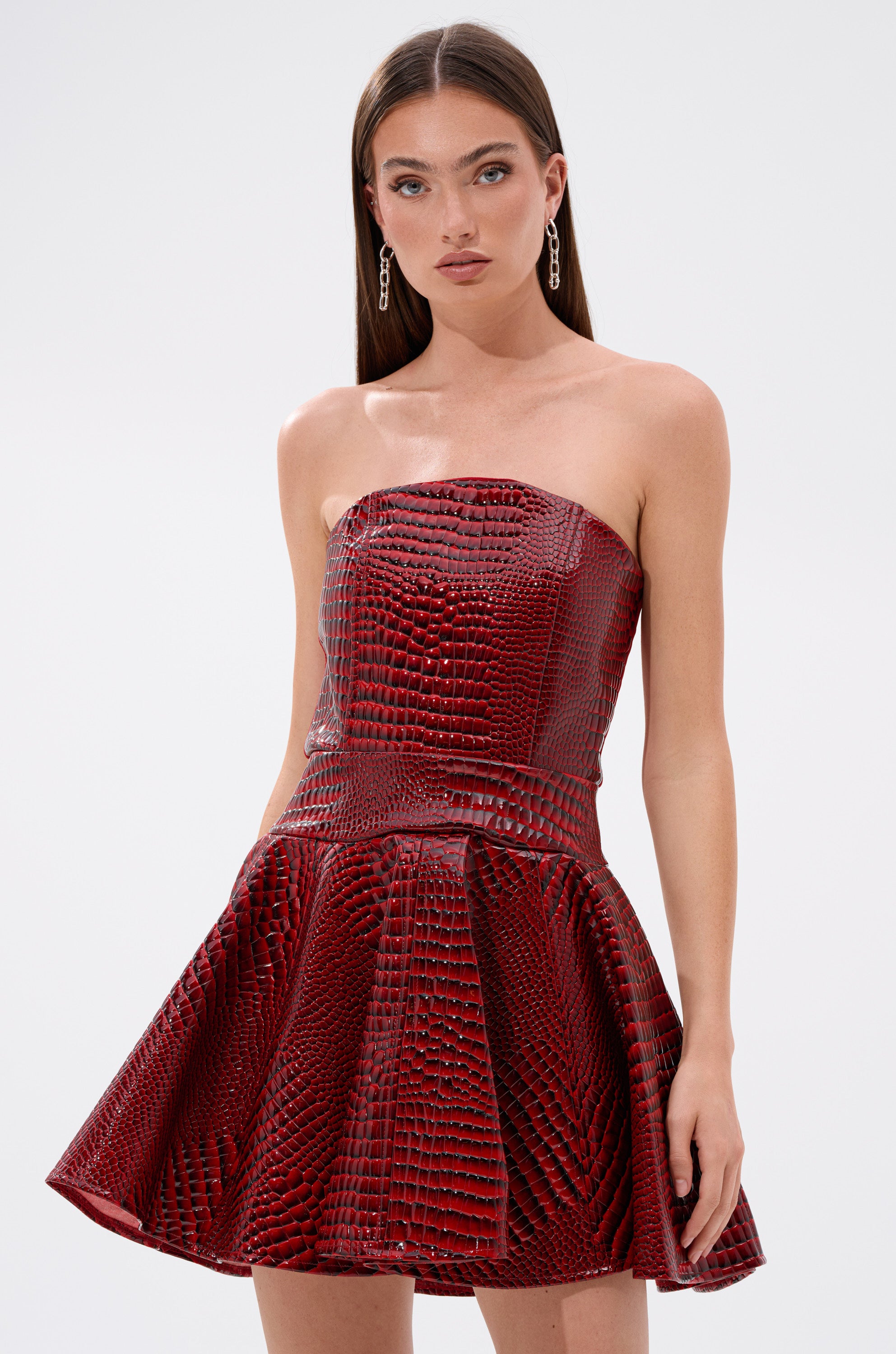 A woman in the VAL PU MINI DRESS IN CROC PRINT, featuring a striking red hue and a corset-style bodice.