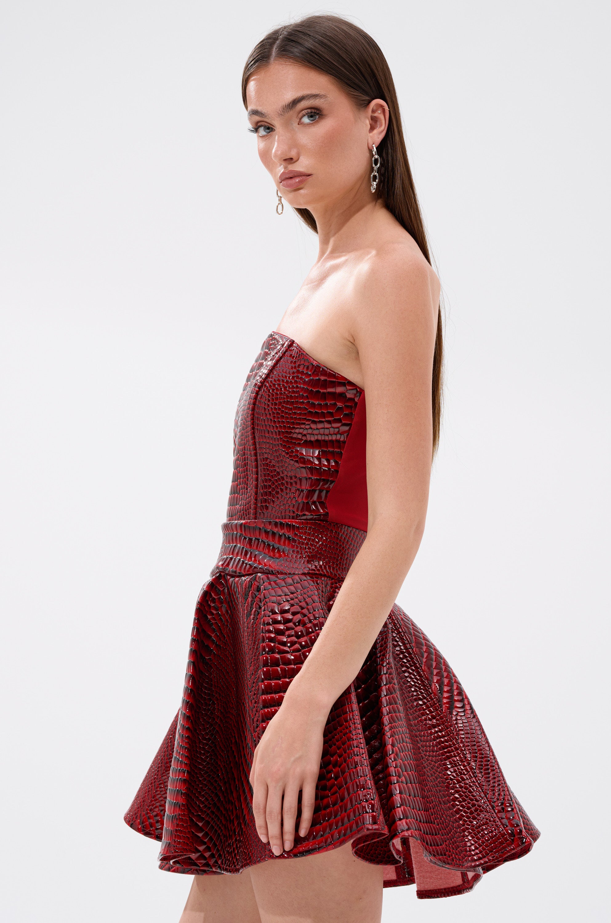 A woman with long brown hair wears the VAL PU MINI DRESS IN CROC PRINT, a strapless shiny red mini dress with a flared skirt and corset-style bodice. She stands in profile against a light background, styled with dangling earrings.