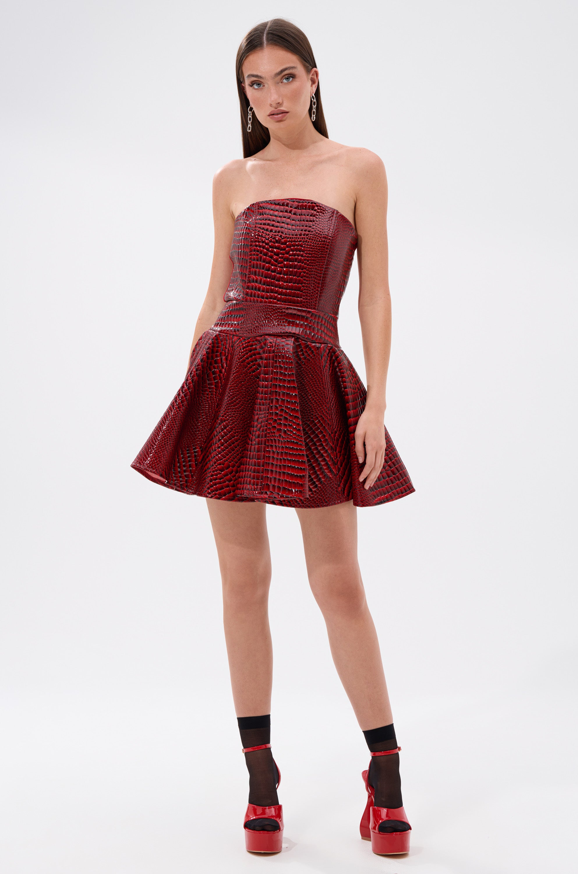 A woman poses against a plain background in the VAL PU MINI DRESS IN CROC PRINT, a shiny red strapless mini dress styled with black ankle socks and bold red platform heels.