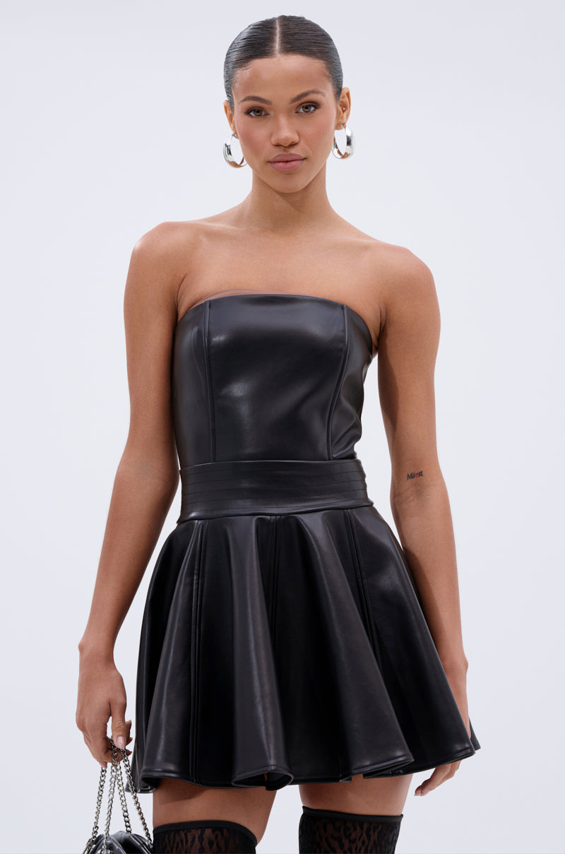 A woman in the VAL PU MINI DRESS—strapless faux leather with a flared skater skirt—paired with large hoop earrings and a chain-strap bag, stands against a plain light background.