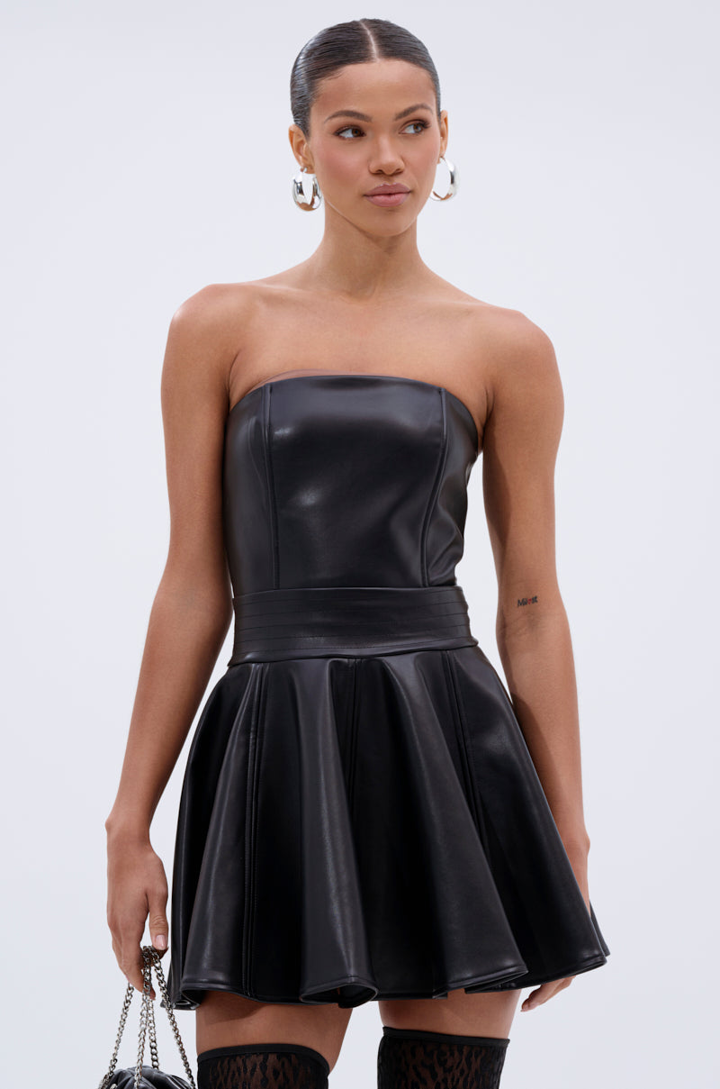 A woman in the VAL PU MINI DRESS—strapless faux leather with a fitted bodice and flared skater skirt—holds a chain-strap bag, sports hoop earrings, and wears her hair in a sleek ponytail as she looks to the side.