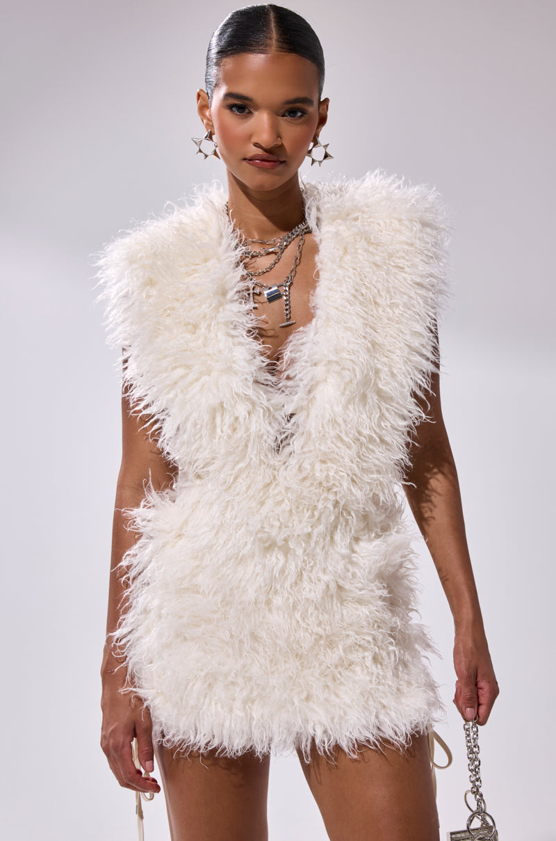 A woman poses confidently against a plain light background in the CALL ME CRAZY FUR MINI DRESS, a sleeveless mini dress with a plunging V-neckline, styled with layered necklaces, earrings, and a small purse.