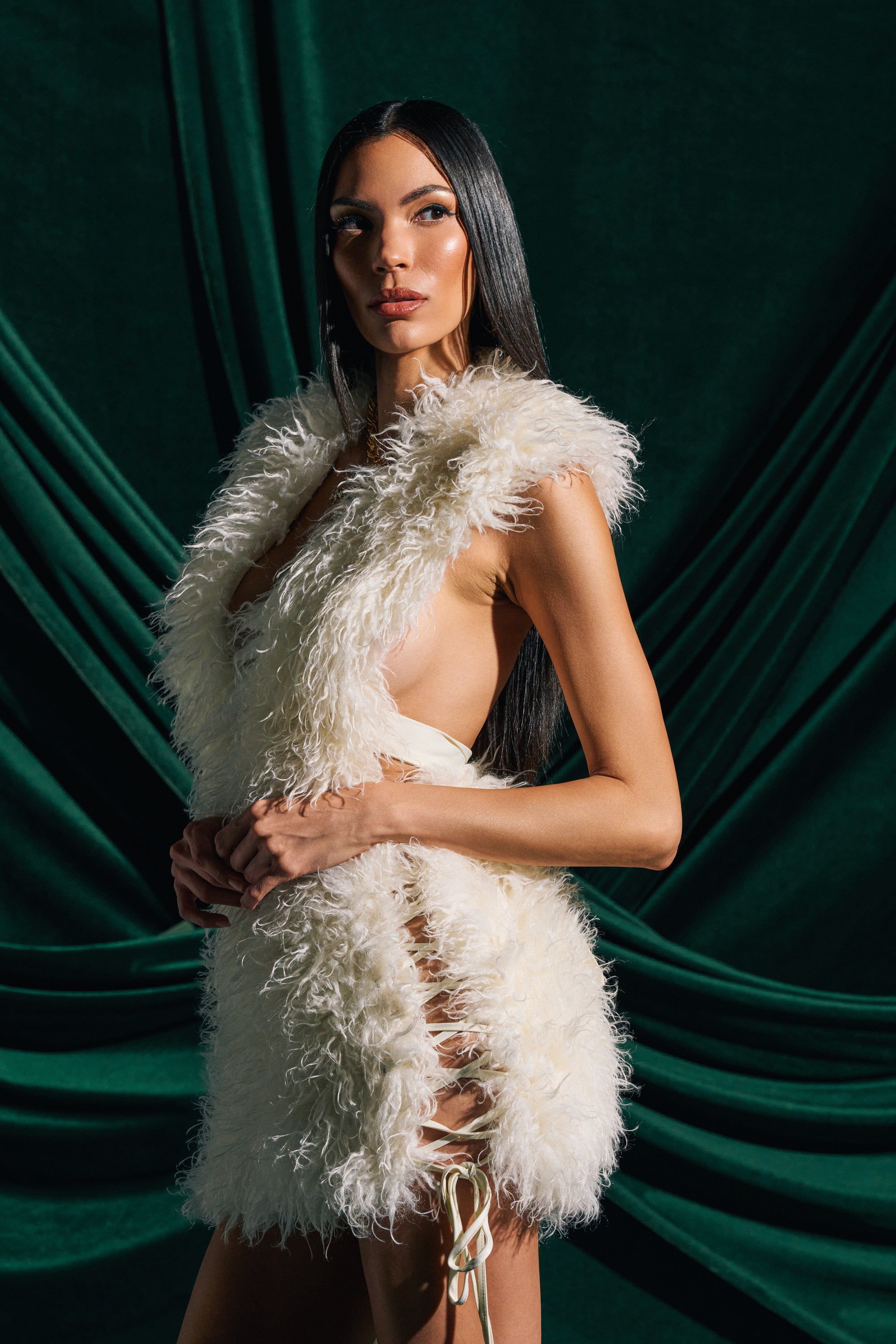 A woman with long dark hair poses before dark green drapery, wearing the CALL ME CRAZY FUR MINI DRESS—a sleeveless faux fur mini with lace-up side seams revealing her sides and back, its texture enhanced by dramatic lighting.