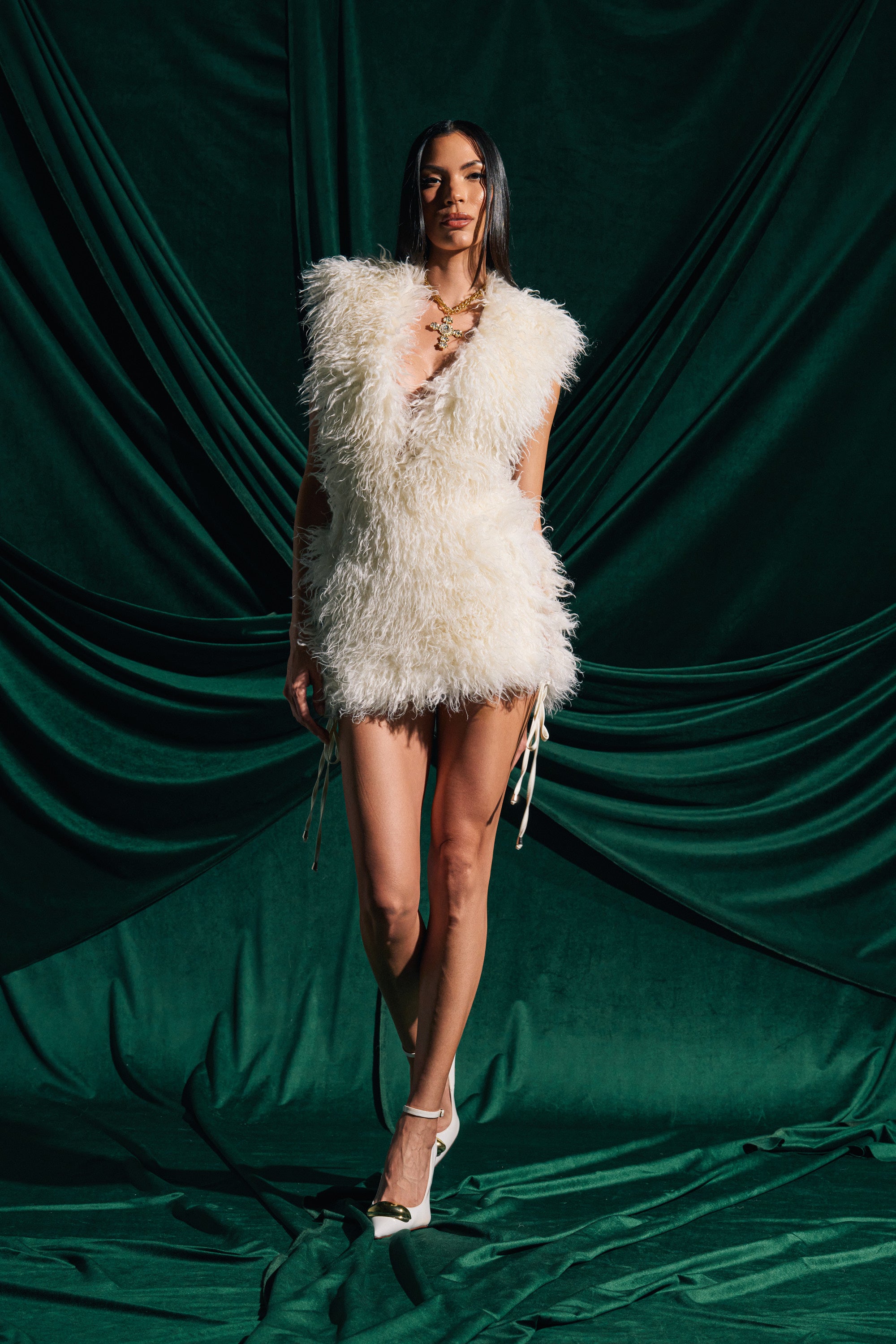 Wearing the CALL ME CRAZY FUR MINI DRESS, a sleeveless white faux fur mini, paired with white heels and a gold necklace, this model stands confidently before emerald-green draped fabric, her long straight dark hair completing the look.
