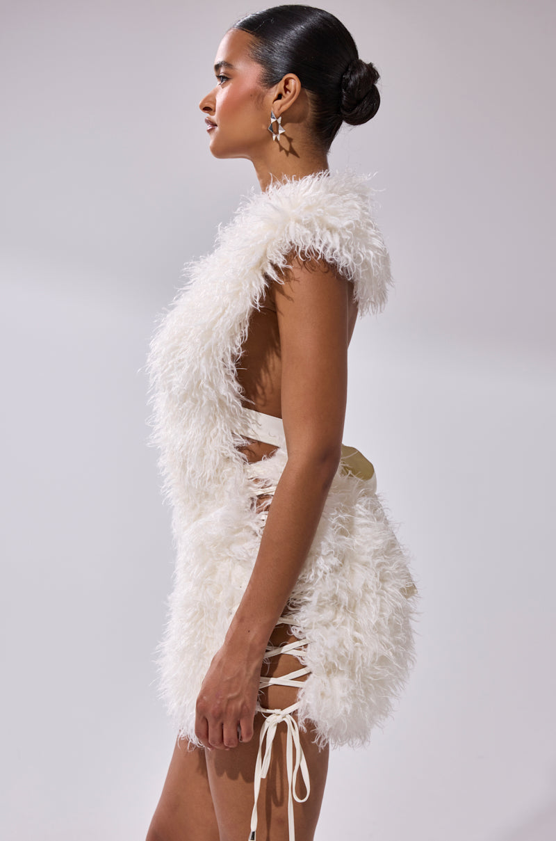 A woman in profile wears the CALL ME CRAZY FUR MINI DRESS—a sleeveless, white faux fur mini with open sides and lace-up ribbons. She has a sleek bun and large hoop earrings, standing against a plain light background.