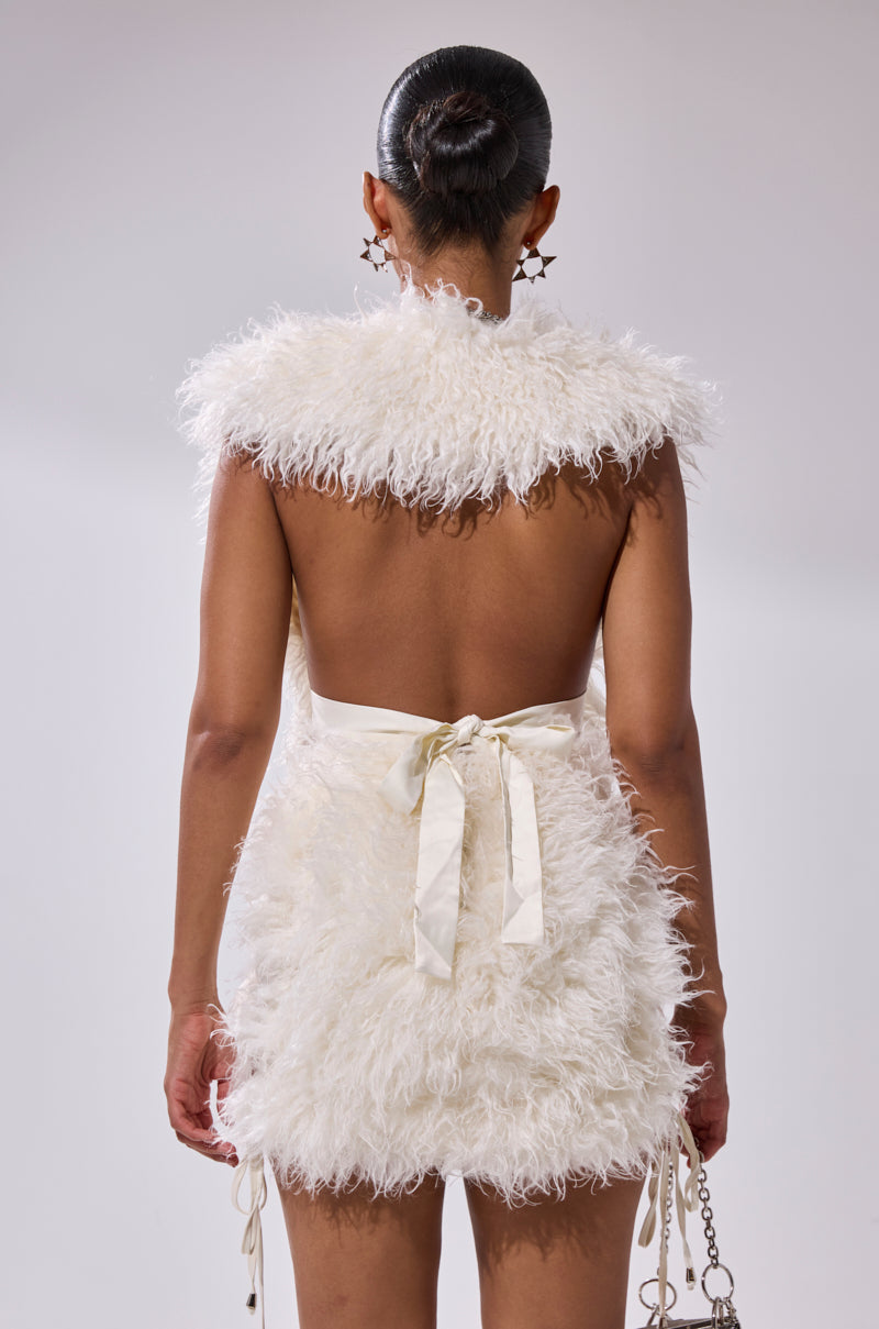 A woman with her hair in a sleek bun wears the CALL ME CRAZY FUR MINI DRESS—white, open-back, faux fur with a satin bow at the waist—holding a small chain purse and standing against a plain light background, facing away.