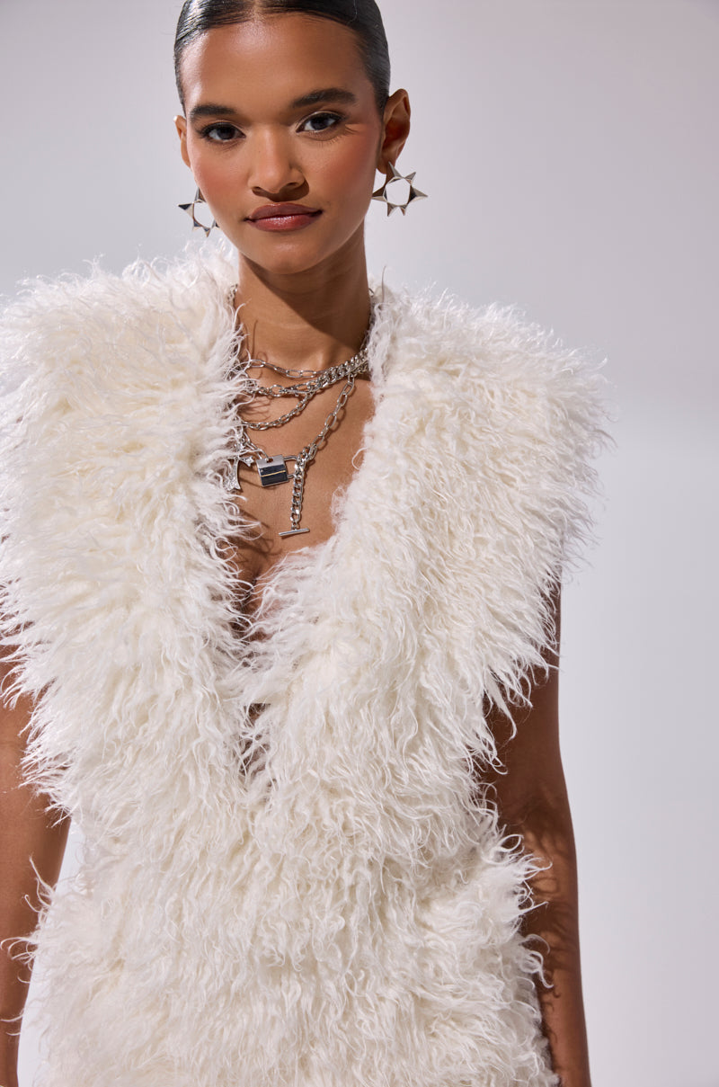 A person with slicked-back hair wears star earrings, layered silver necklaces, and the CALL ME CRAZY FUR MINI DRESS—a sleeveless, fluffy white faux fur mini dress with a plunging V-neckline—against a plain light background.