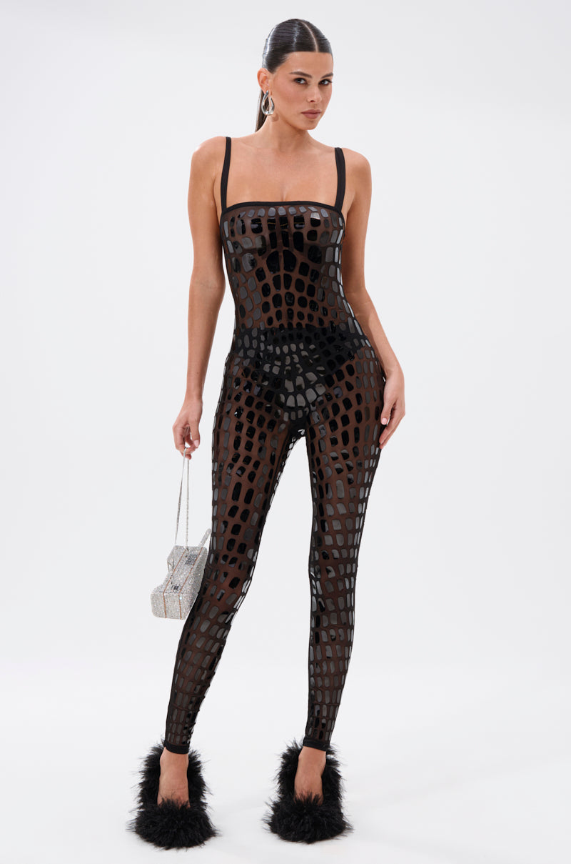 A woman poses confidently in the BLING BLOW MESH JUMPSUIT—a sheer black jumpsuit with abstract cutouts—paired with black undergarments, fluffy black heels, and a silver handbag against a white background for a high-fashion look.