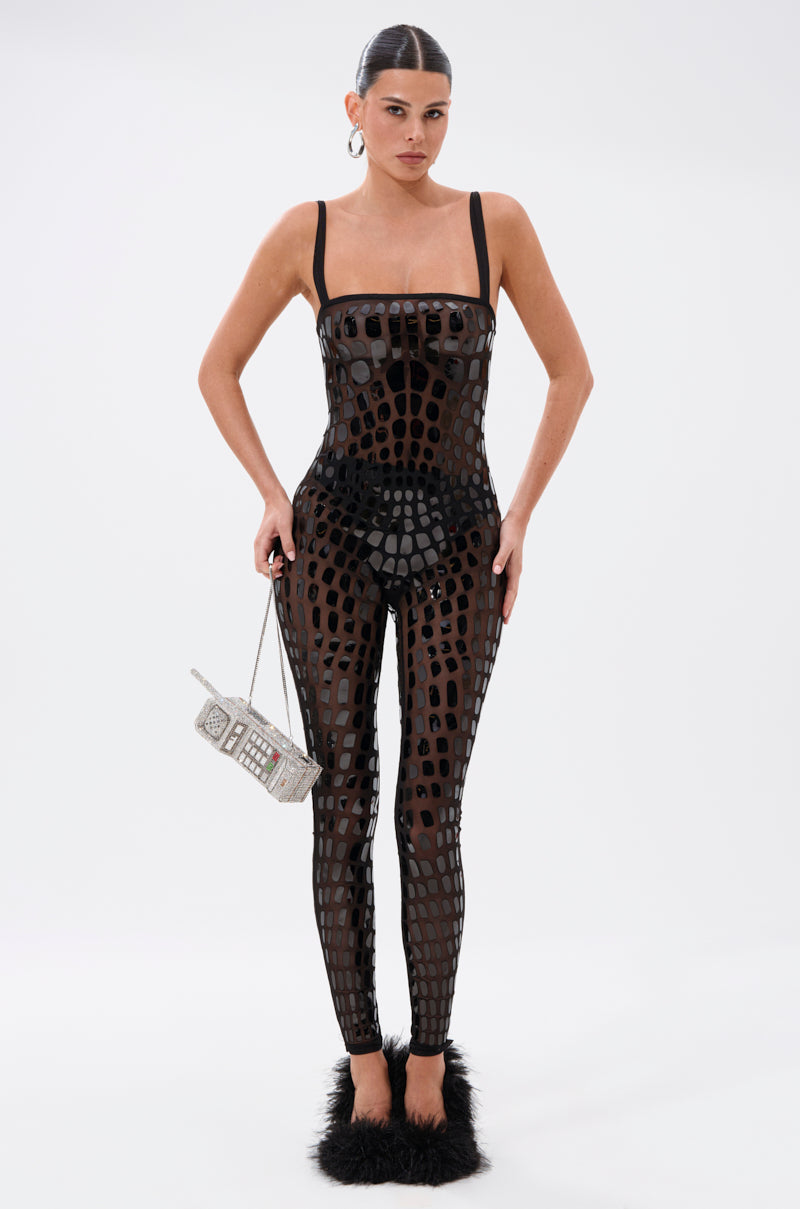 A woman with slicked-back hair and large hoops wears the BLING BLOW MESH JUMPSUIT, a black sheer geometric-patterned outfit, paired with black furry shoes and a retro cellphone-shaped beaded handbag against a plain white background.