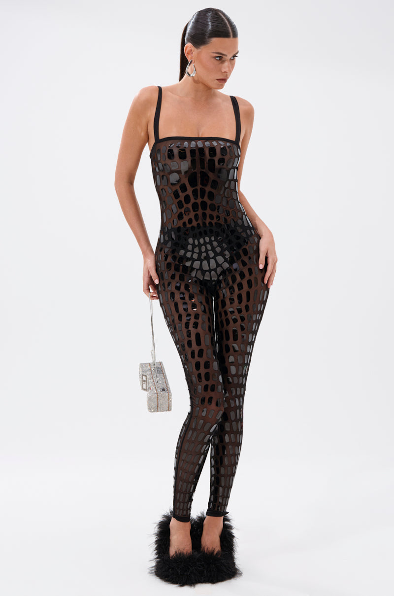 A woman models the BLING BLOW MESH JUMPSUIT, featuring geometric cutout patterns, styled with black undergarments, fuzzy black slippers, and a small silver handbag against a plain white background.