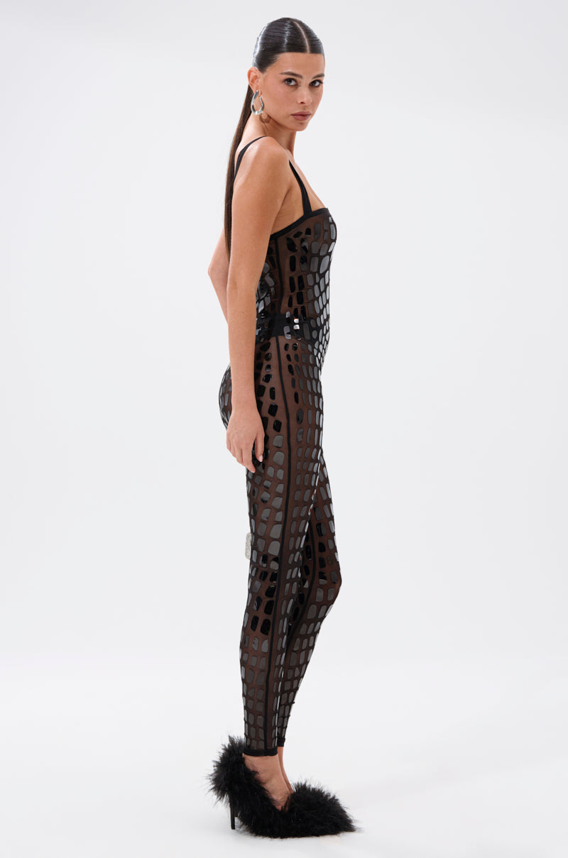 A woman poses sideways in the BLING BLOW MESH JUMPSUIT, a sheer black design with geometric embellishments, paired with black feather-detailed heels and sleek straight hair against a plain white background.