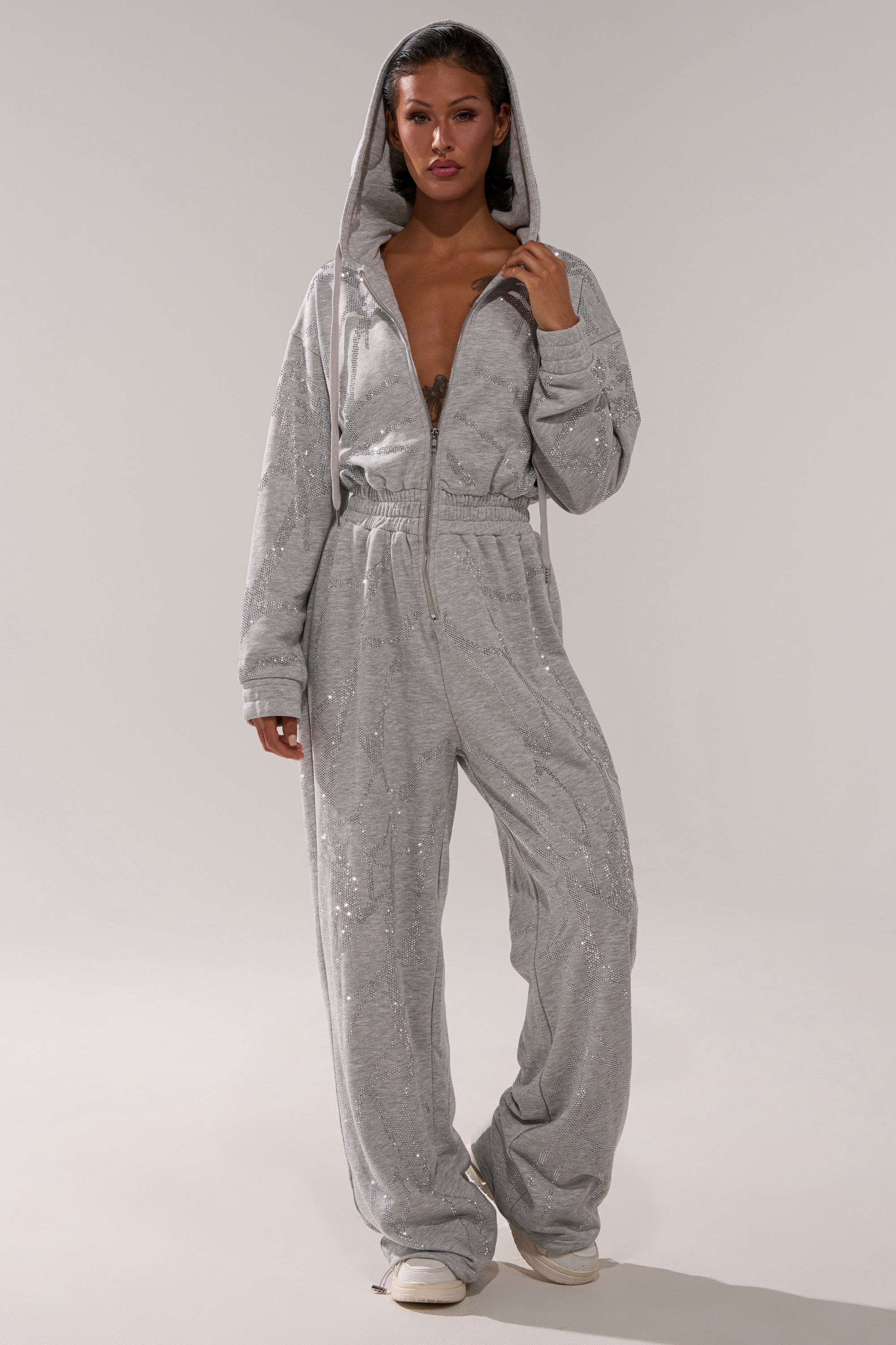 A person wearing the NEVER BUGGIN PRINTED FRENCH TERRY JUMPSUIT with rhinestone details and a front zipper, slightly open at the chest, paired with white shoes, stands against a plain background.