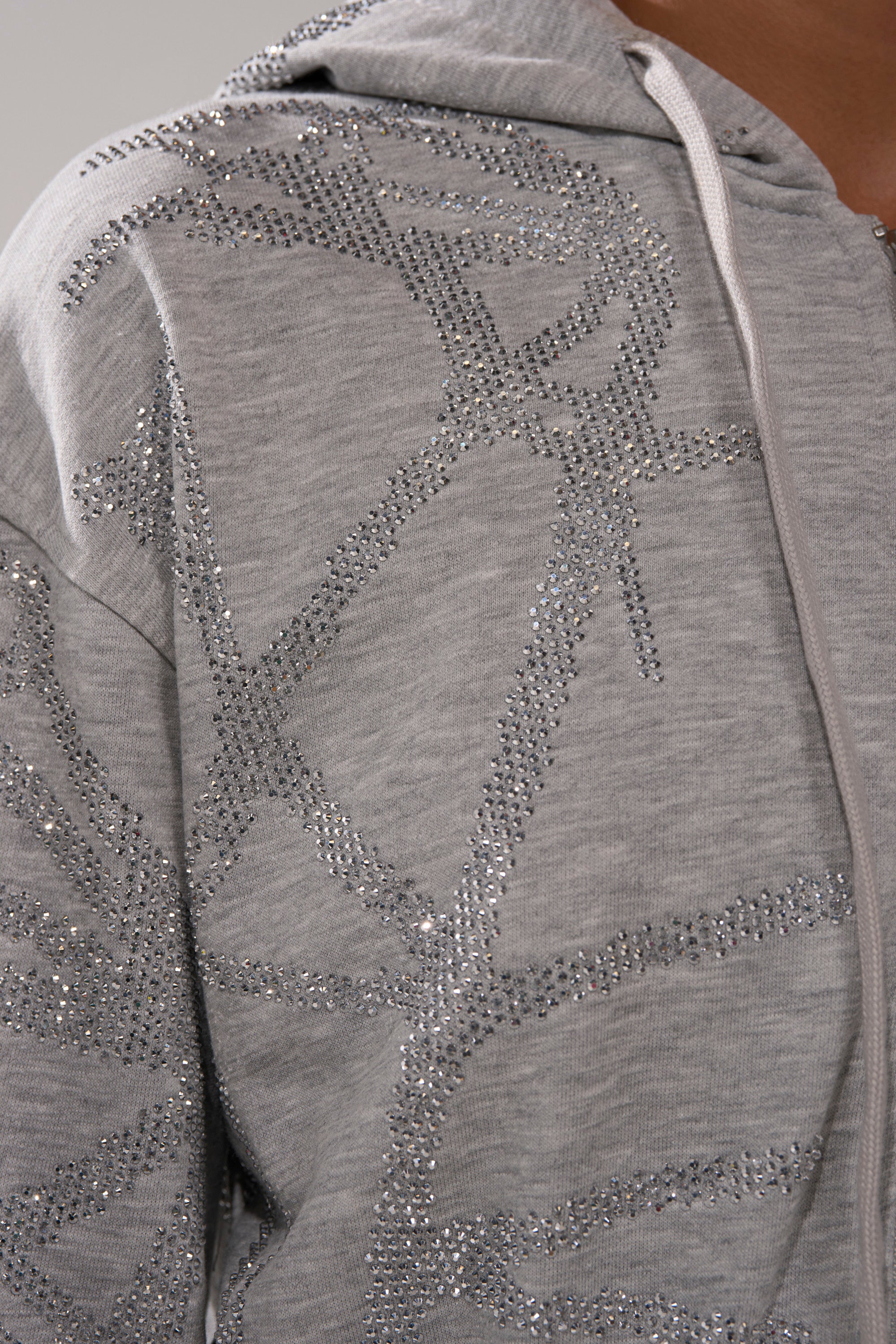 Close-up of a person wearing the NEVER BUGGIN PRINTED FRENCH TERRY JUMPSUIT in light gray, featuring sparkling rhinestone accents and a white zipper. Abstract rhinestone patterns add shimmer to this modern hooded one-piece.