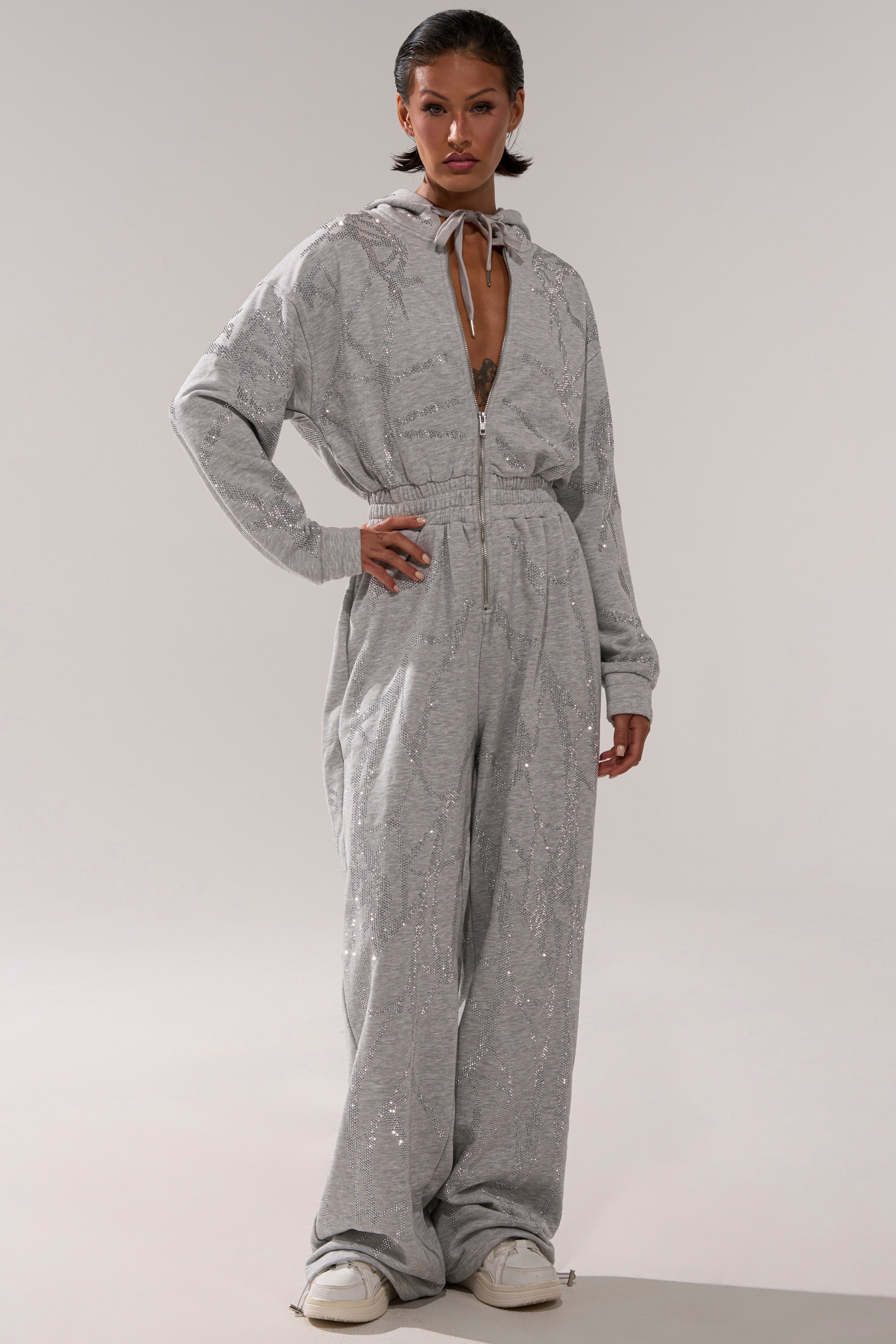 A person stands confidently in the NEVER BUGGIN PRINTED FRENCH TERRY JUMPSUIT, a loose-fitting, light gray one-piece with a zipper front, wide legs, and white sneakers. The outfit features a subtle shimmer against a plain background.