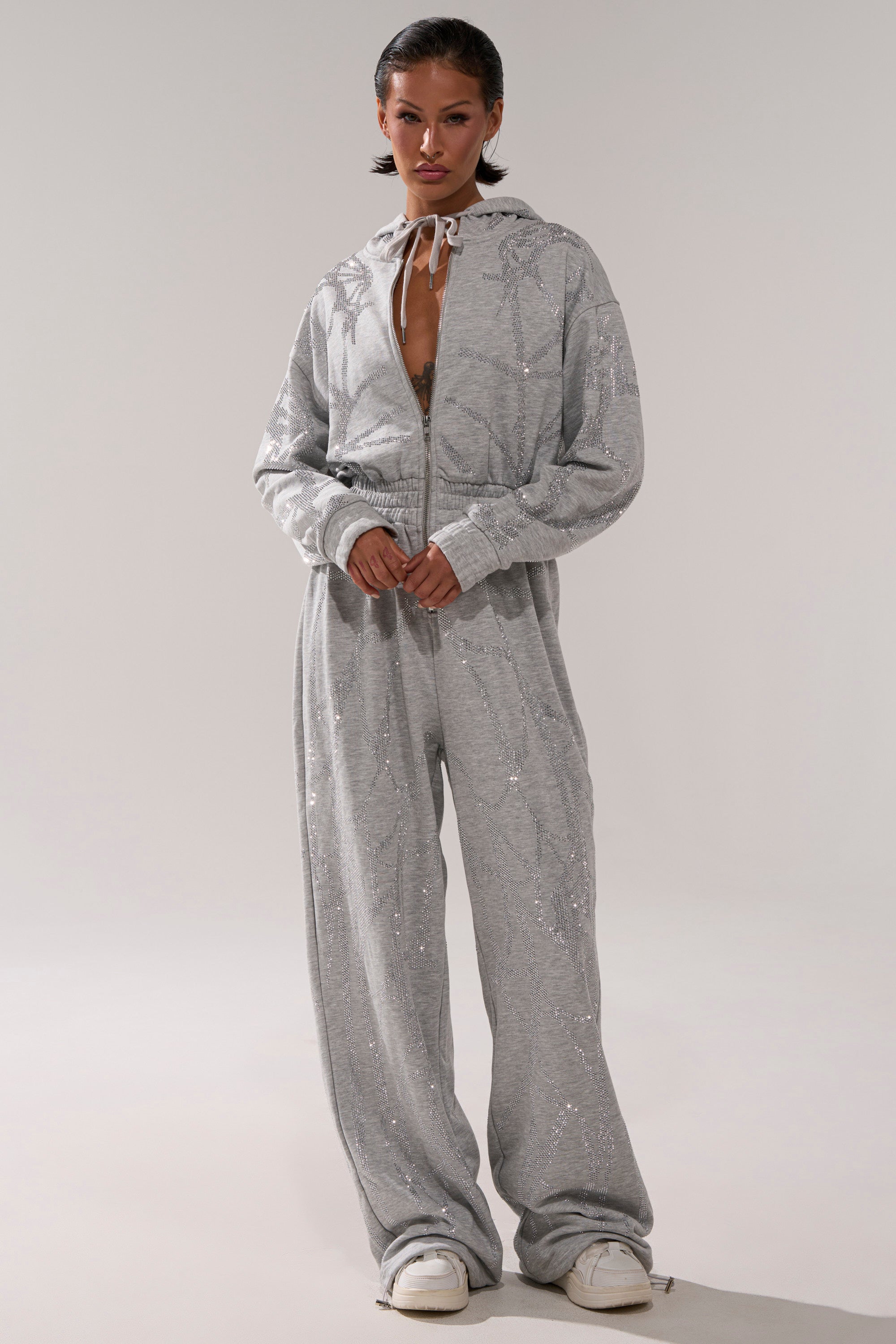 A person stands against a plain background wearing the AKIRA Label NEVER BUGGIN Printed French Terry Jumpsuit with a relaxed, comfortable fit, paired with white sneakers.