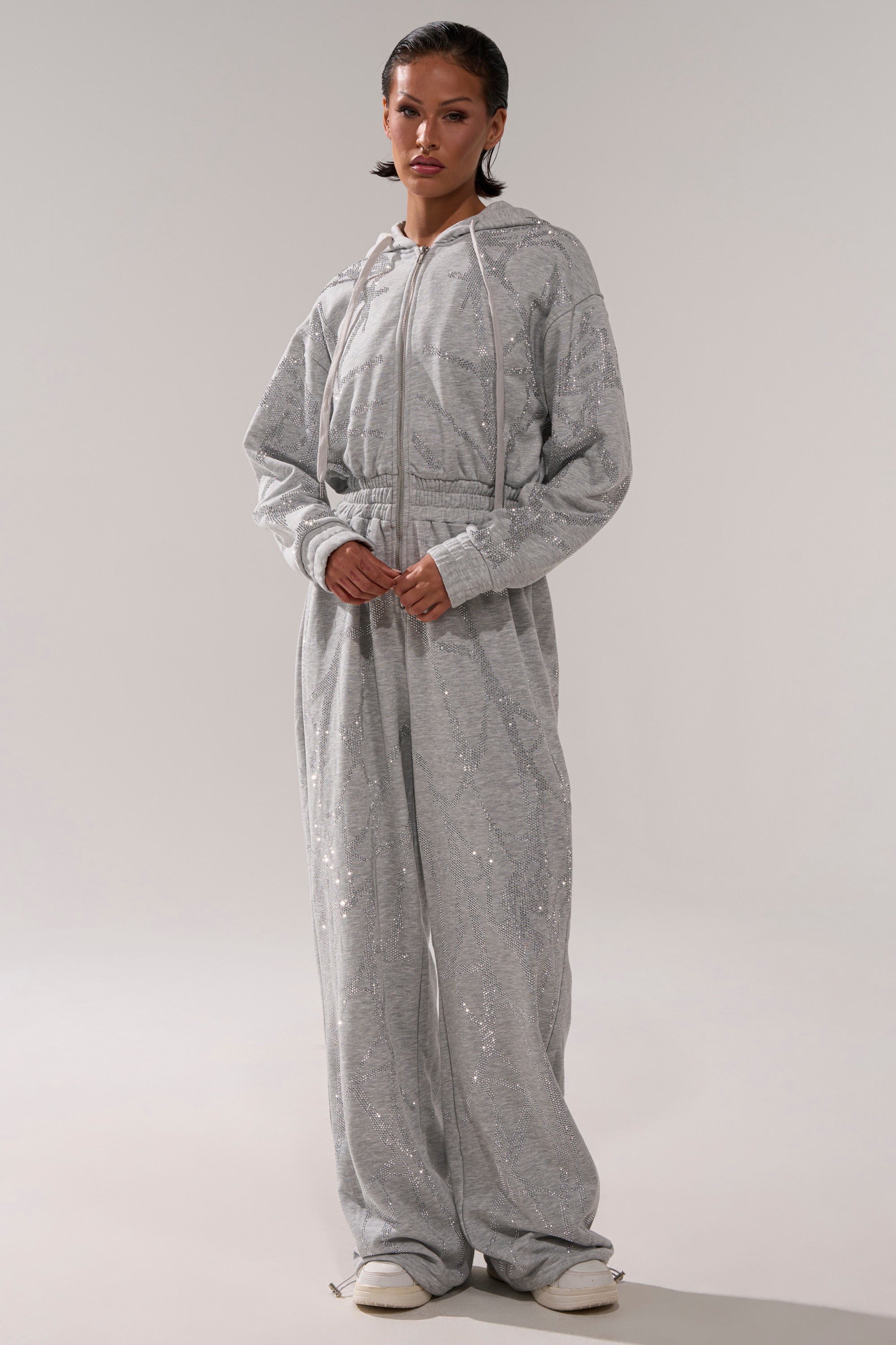 A person faces forward, wearing the NEVER BUGGIN PRINTED FRENCH TERRY JUMPSUIT in light gray with subtle rhinestone details. They have short dark hair, white shoes, and stand against a plain, light background.