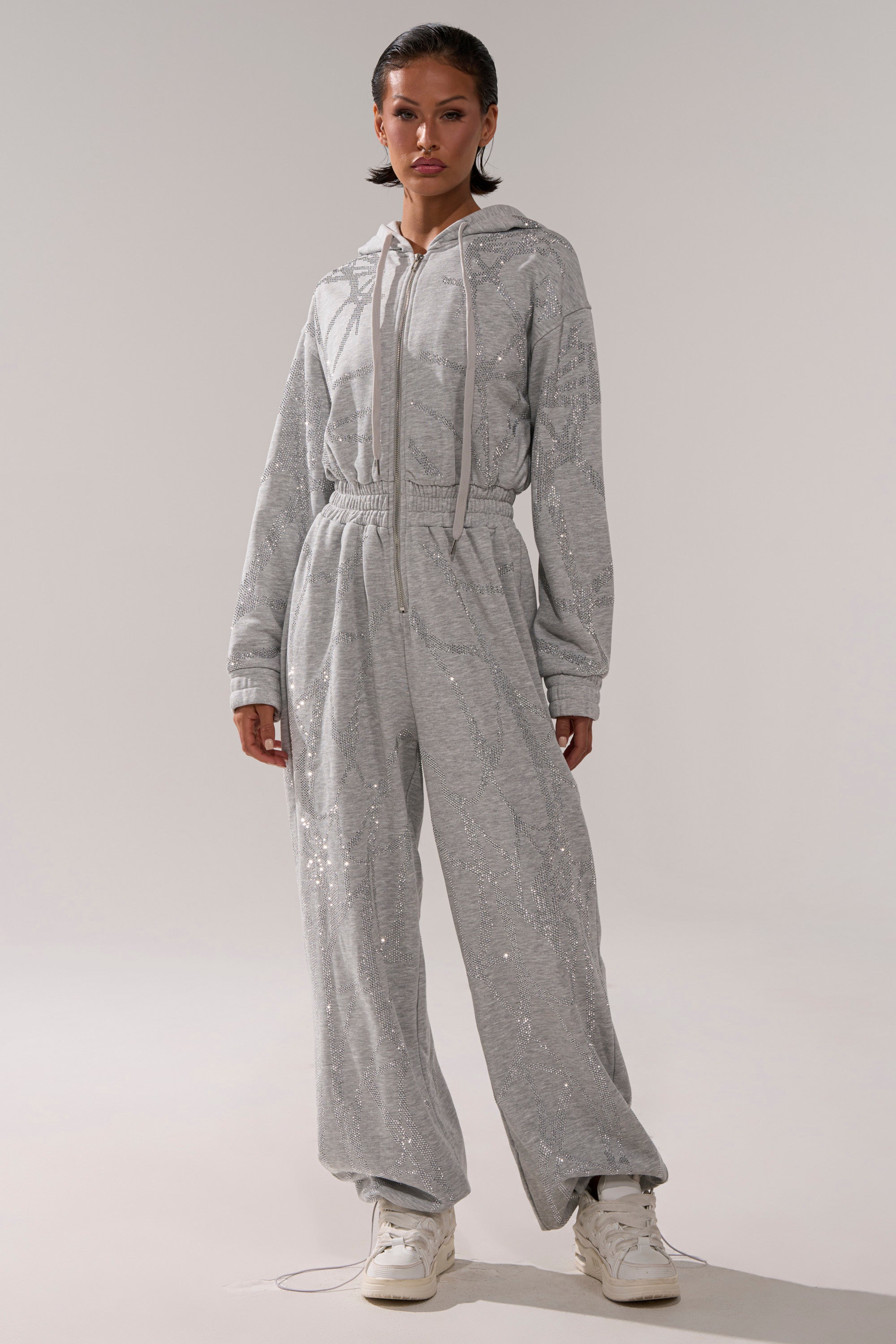 A woman wears the NEVER BUGGIN PRINTED FRENCH TERRY JUMPSUIT with rhinestone accents and an elastic waistband, paired with white sneakers, standing against a plain light background.