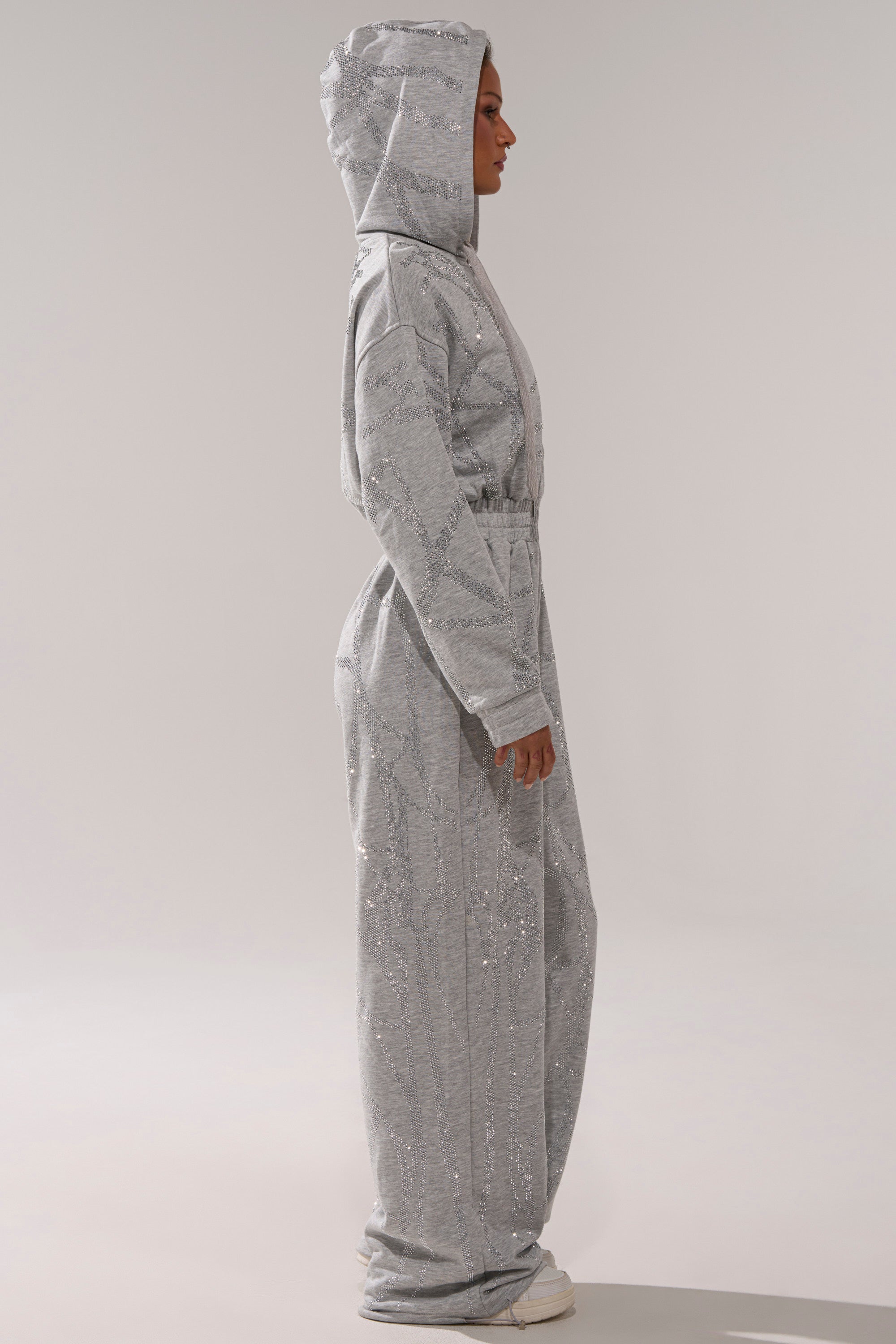 Wearing the NEVER BUGGIN PRINTED FRENCH TERRY JUMPSUIT, a person stands in profile against a plain background—showcasing its rhinestone details, light gray hue, long sleeves, and full-length pants paired with white sneakers for modern loungewear flair.