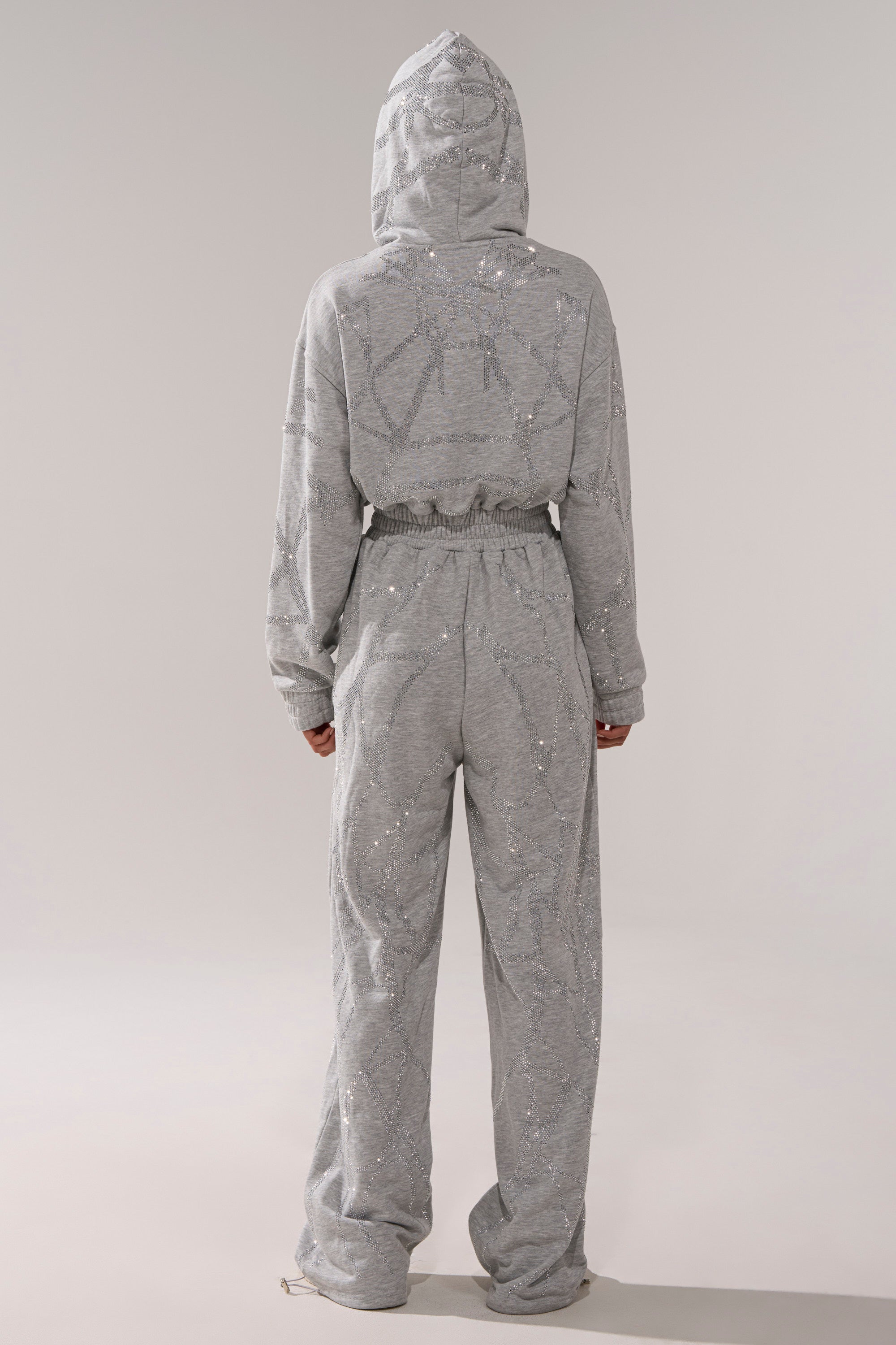 A person wearing the NEVER BUGGIN PRINTED FRENCH TERRY JUMPSUIT with rhinestone details, a hood up, and a relaxed fit stands facing away from the camera against a plain background.