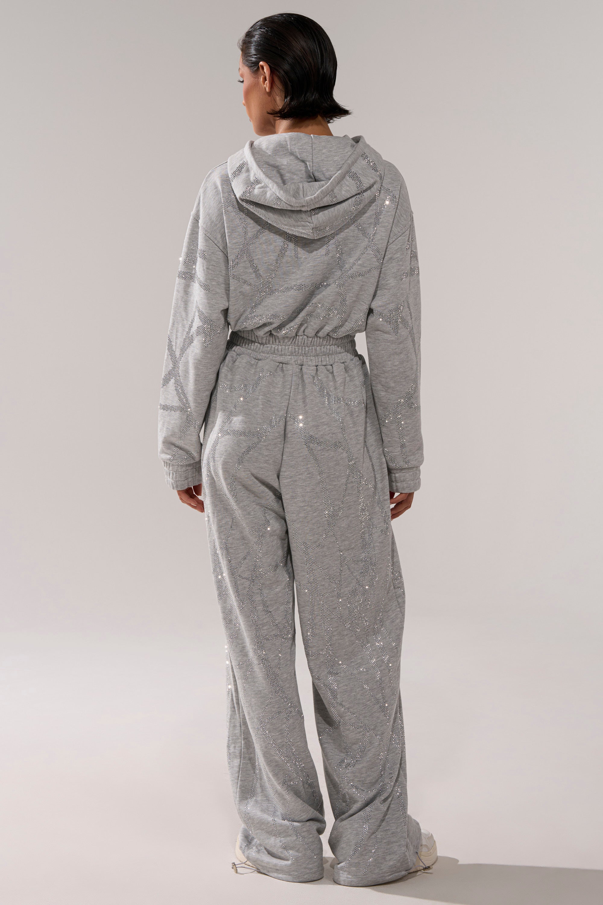 A person stands facing away, wearing the loose-fitting, light gray NEVER BUGGIN PRINTED FRENCH TERRY JUMPSUIT with subtle rhinestone embellishments. The background is plain and light-colored.