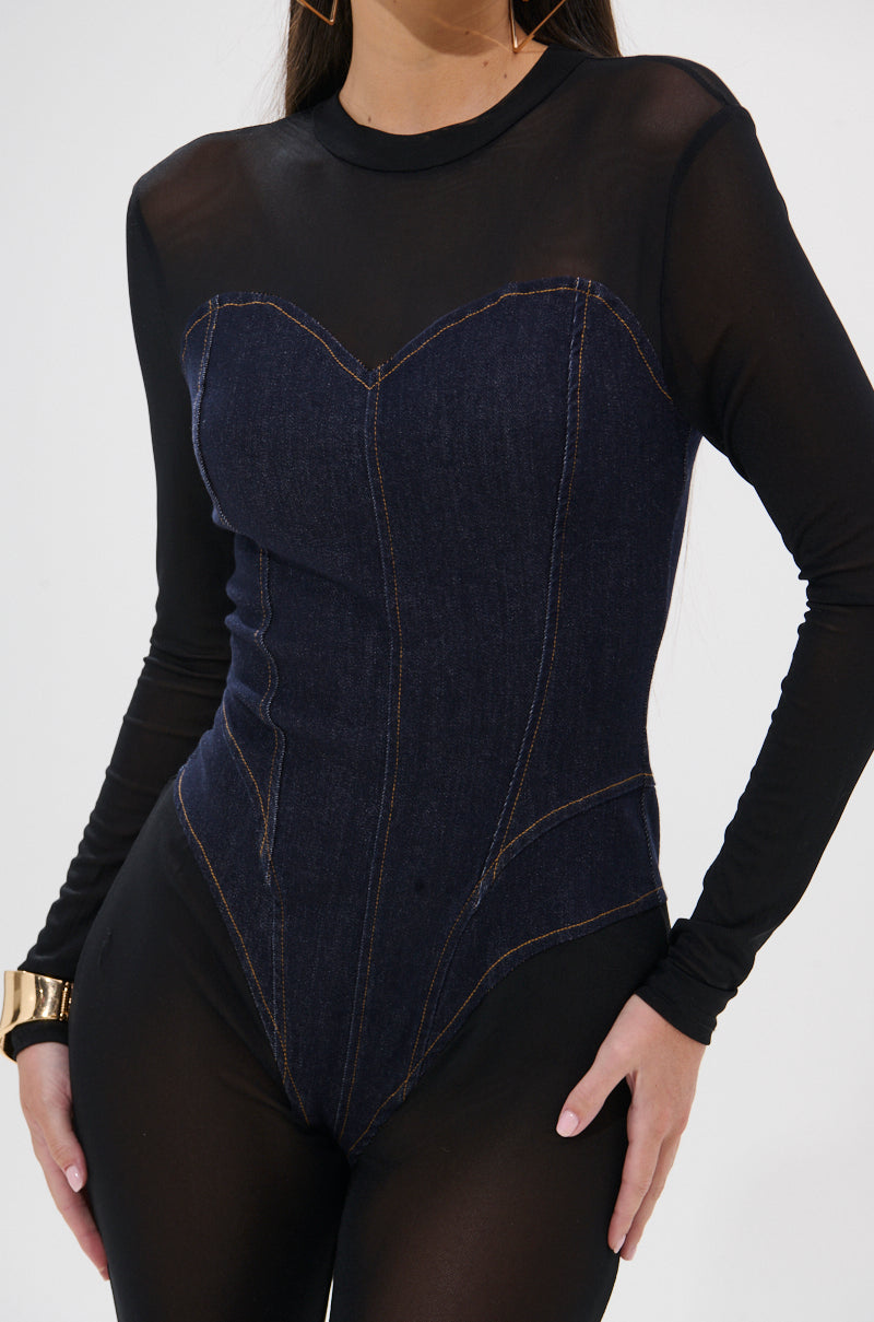 A woman wears the NEW YEAR SAME ME MESH DENIM JUMPSUIT, featuring a sheer black long-sleeve top with a curve-hugging fit and dark denim corset bodice. She accessorizes with gold hoop earrings and a chunky gold bracelet. Cropped shoulder to mid-thigh.