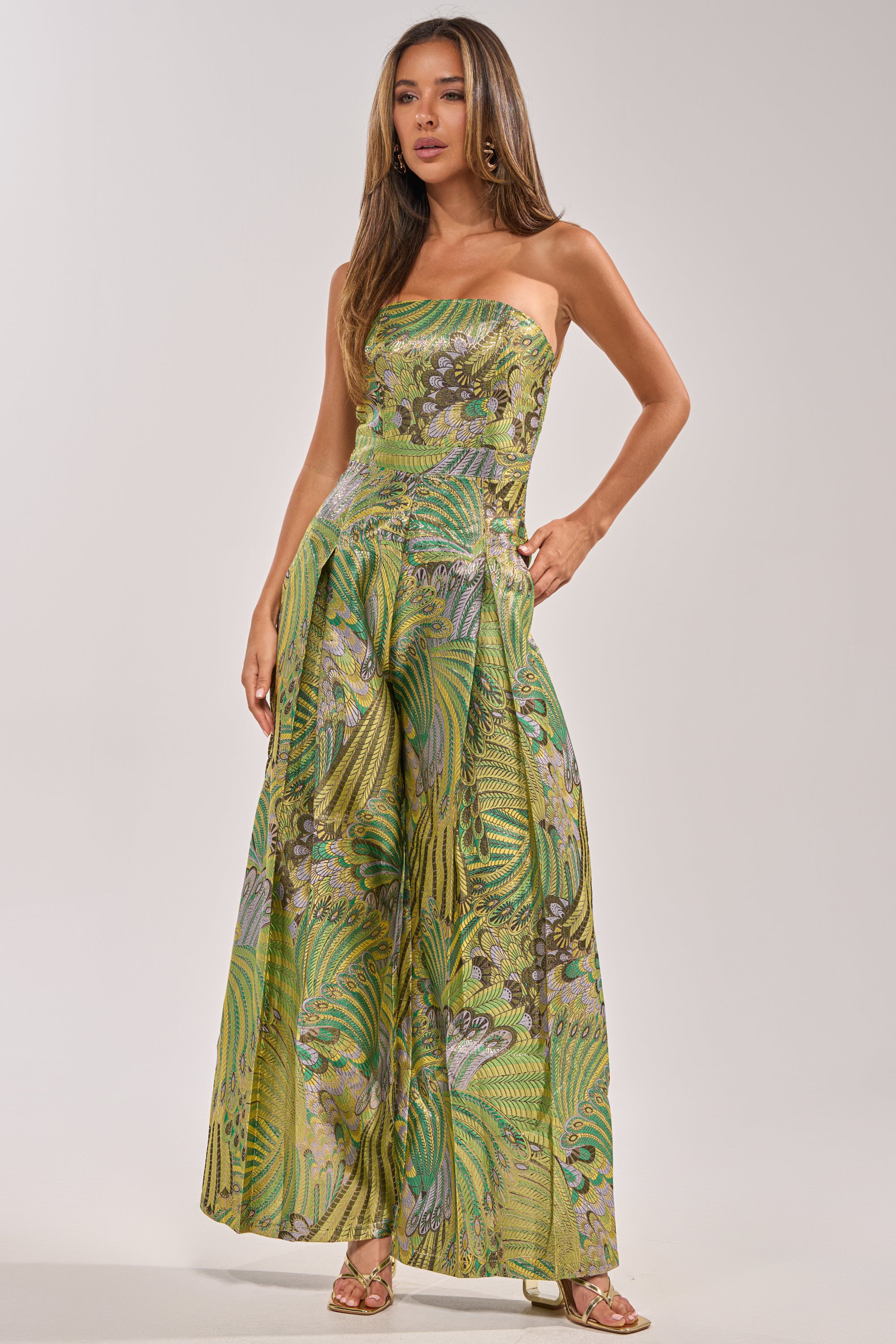 A woman poses in the NO ONE LIKE ME PRINTED JUMPSUIT IN GREEN, a strapless wide-leg jumpsuit with a vibrant botanical print, styled with gold hoop earrings and sandals against a plain light background for a chic look.