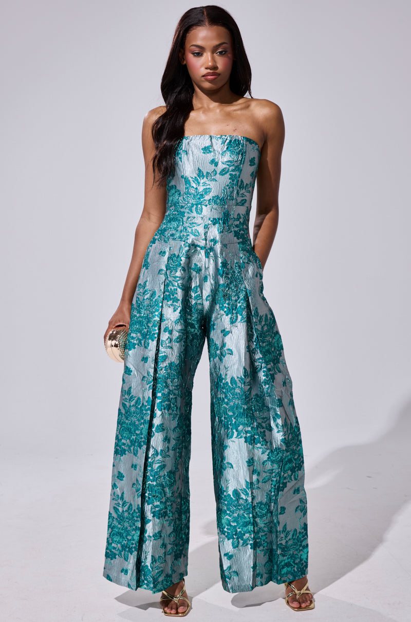 A woman wears the NO ONE LIKE ME PRINTED JUMPSUIT, a strapless, wide-leg style in teal and silver brocade. She accessorizes with a small clutch and gold open-toe heels, posing against a plain light gray background.