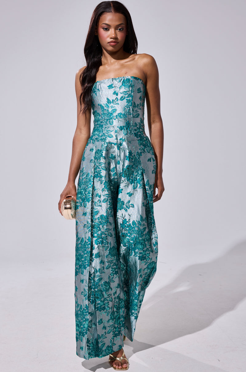 A woman poses confidently against a light gray background wearing the NO ONE LIKE ME PRINTED JUMPSUIT, a strapless wide-leg piece in teal and silver floral print, styled with a small clutch and gold sandals.