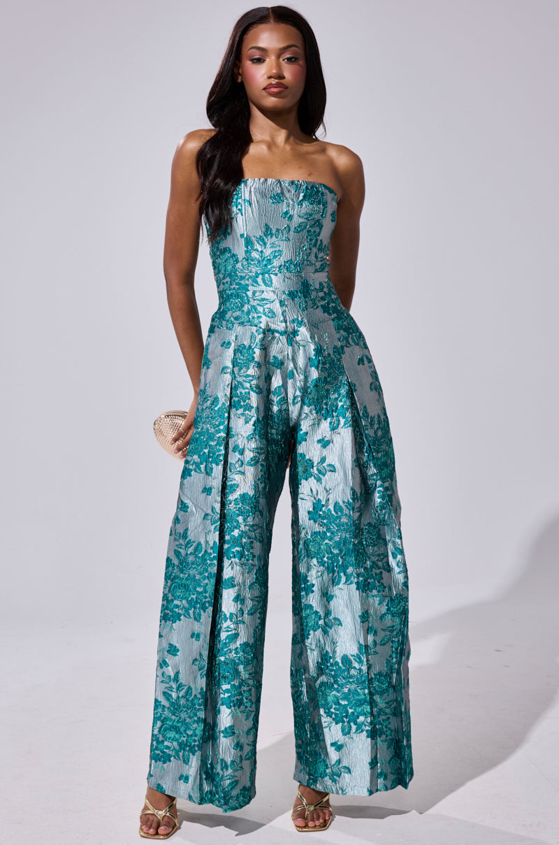 A woman poses against a light background in the NO ONE LIKE ME PRINTED JUMPSUIT, featuring a strapless wide-leg design with teal and silver floral brocade. She accessorizes with a clutch, gold heels, and loose wavy hair.