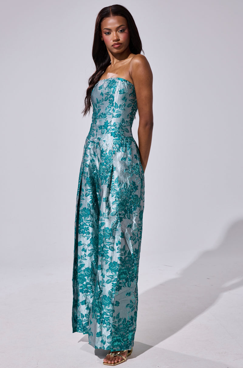 A woman with long dark hair models the NO ONE LIKE ME PRINTED JUMPSUIT, a strapless, floor-length piece featuring a teal floral brocade pattern. She poses with her arms relaxed at her sides against a plain light background.