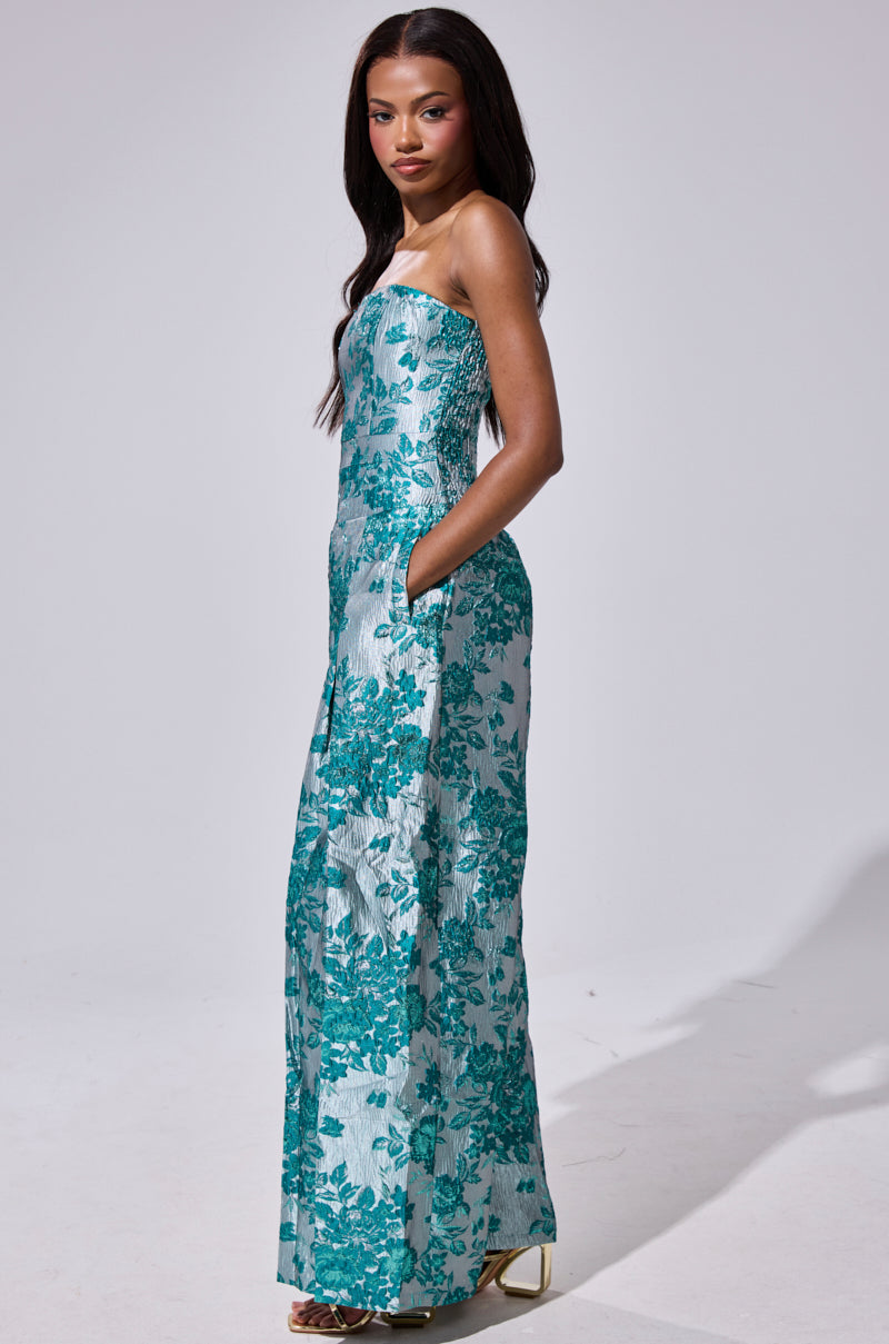 A woman stands in the NO ONE LIKE ME PRINTED JUMPSUIT—a strapless, floor-length silver piece with teal floral patterns and pockets—paired with gold heels, set against a plain light background.