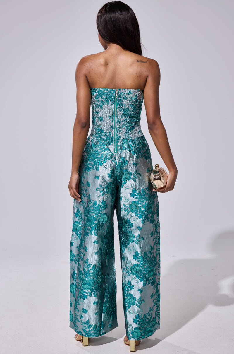 A woman faces away, wearing the NO ONE LIKE ME PRINTED JUMPSUIT—a strapless teal and silver floral brocade wide-leg style. She holds a small clutch in her right hand and lets her long dark hair flow down her back.
