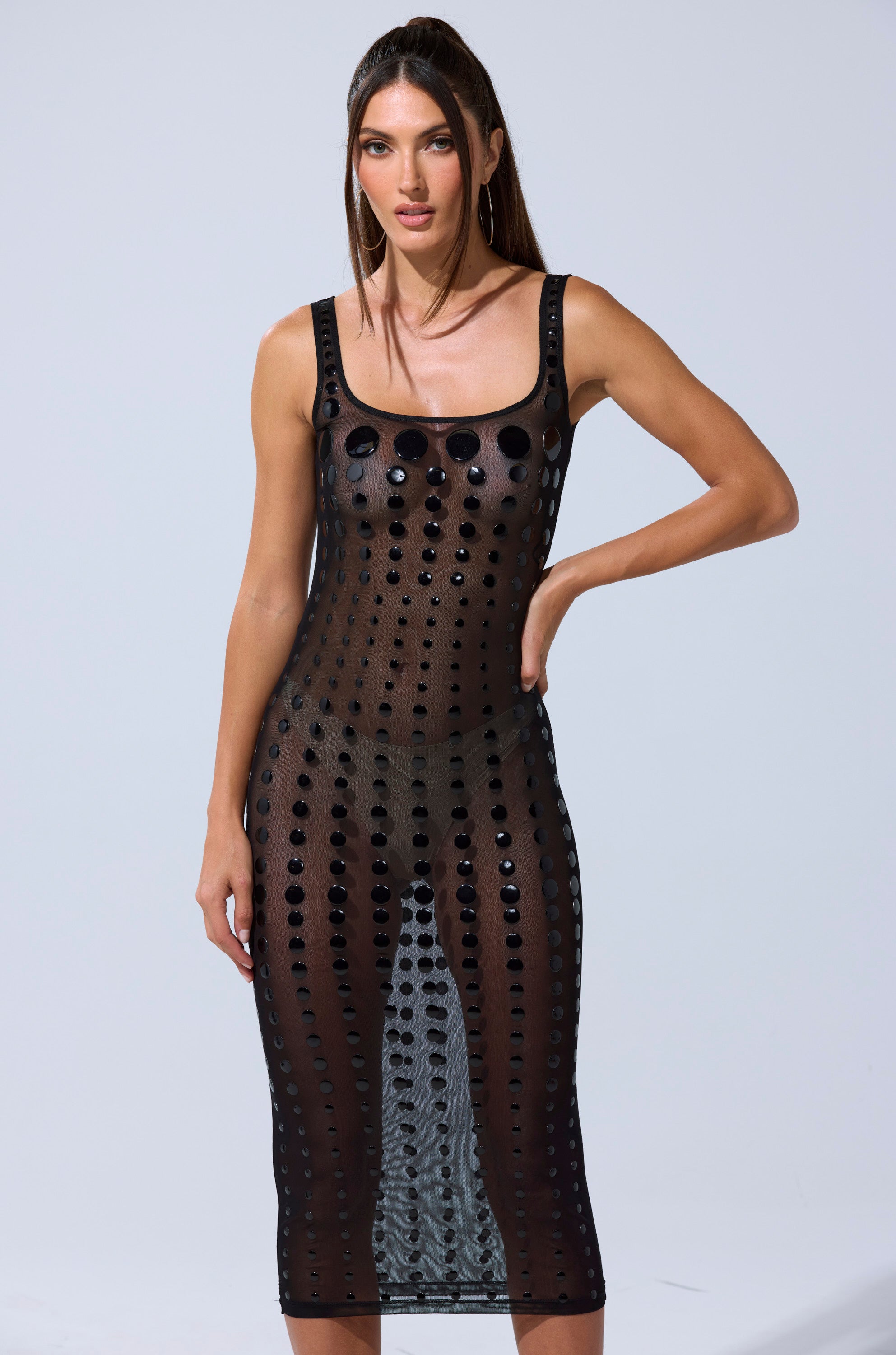 A woman stands confidently in the ROUND OF APPLAUSE MIDI DRESS—a sleeveless, sheer black mesh bodycon midi with a scoop neckline and rows of round black embellishments—set against a plain light background.