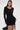 A woman poses in the RUN THIS TOWN LACE UP MINI DRESS, featuring grommets on the sleeves, sides, and front. She pairs the bodycon LBD with a black handbag and wears her long straight hair sleekly against a light background.
