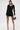 A woman stands confidently in the RUN THIS TOWN LACE UP MINI DRESS, paired with black heels and a silver purse. She wears silver hoop earrings, has straight dark hair, and poses against a plain white background.