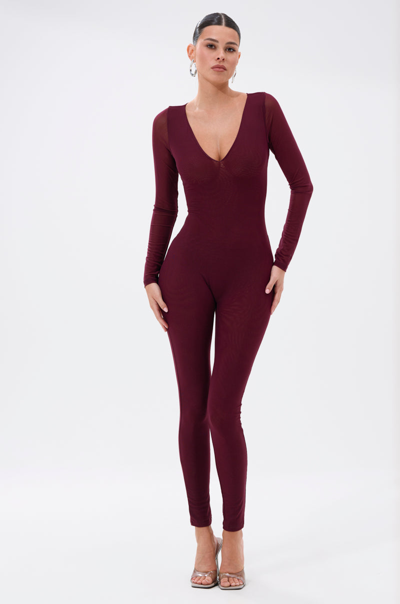 A woman poses confidently against a white background wearing the KEEPIN IT SIMPLE MESH JUMPSUIT IN BURGUNDY, featuring a fitted, long-sleeve, deep V-neck design and paired with silver high-heeled sandals. Her hair is pulled back. 1