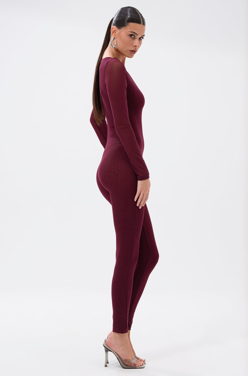 A woman with long, straight hair in a ponytail models the KEEPIN IT SIMPLE MESH JUMPSUIT IN BURGUNDY and clear high heels, standing sideways against a plain white background. 3