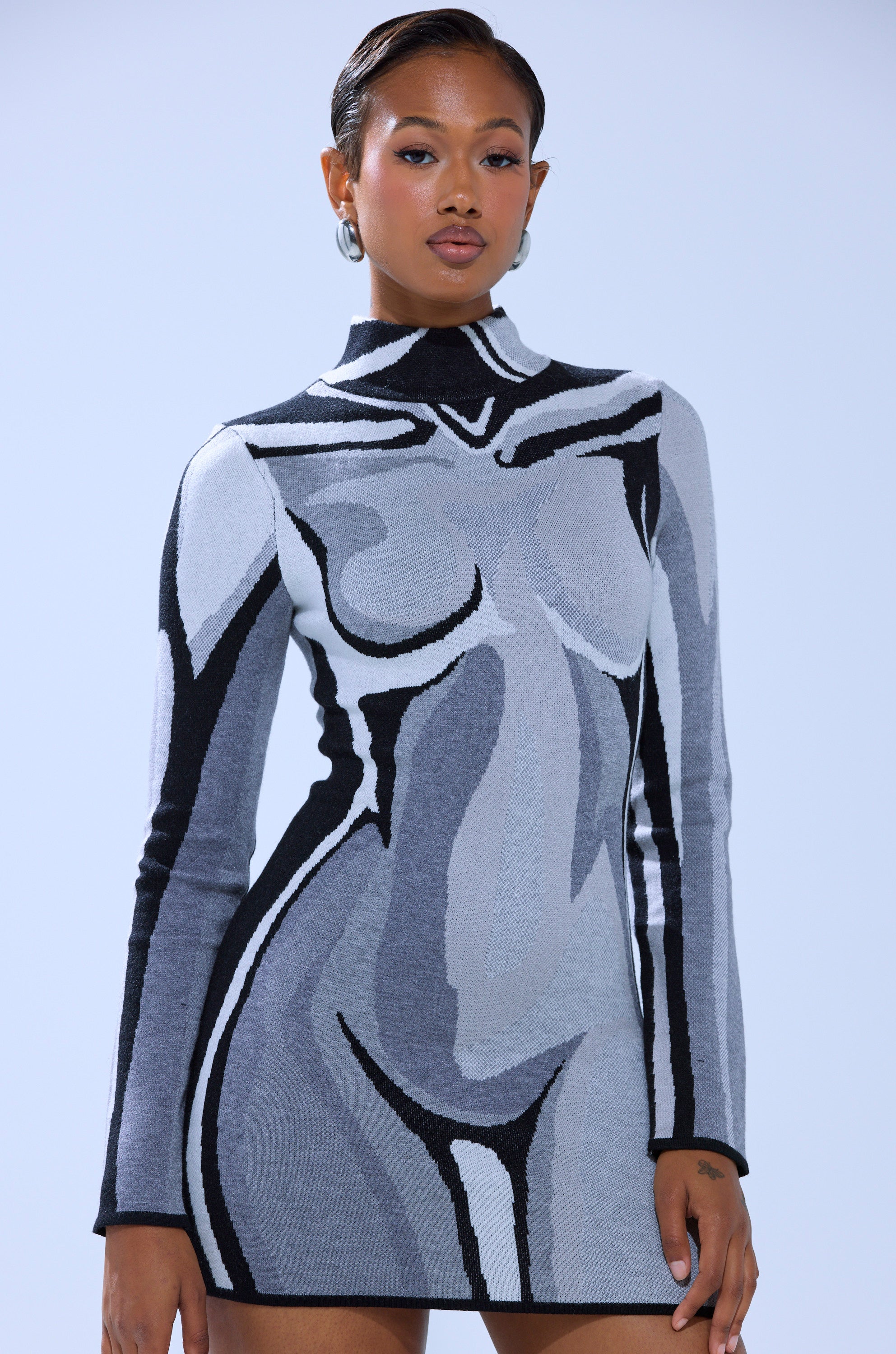 A woman wears the A LIGHT SERVE PRINTED MINI DRESS, a curve-hugging knit with an abstract nude female body design in gray, black, and white, set against a plain light blue background.