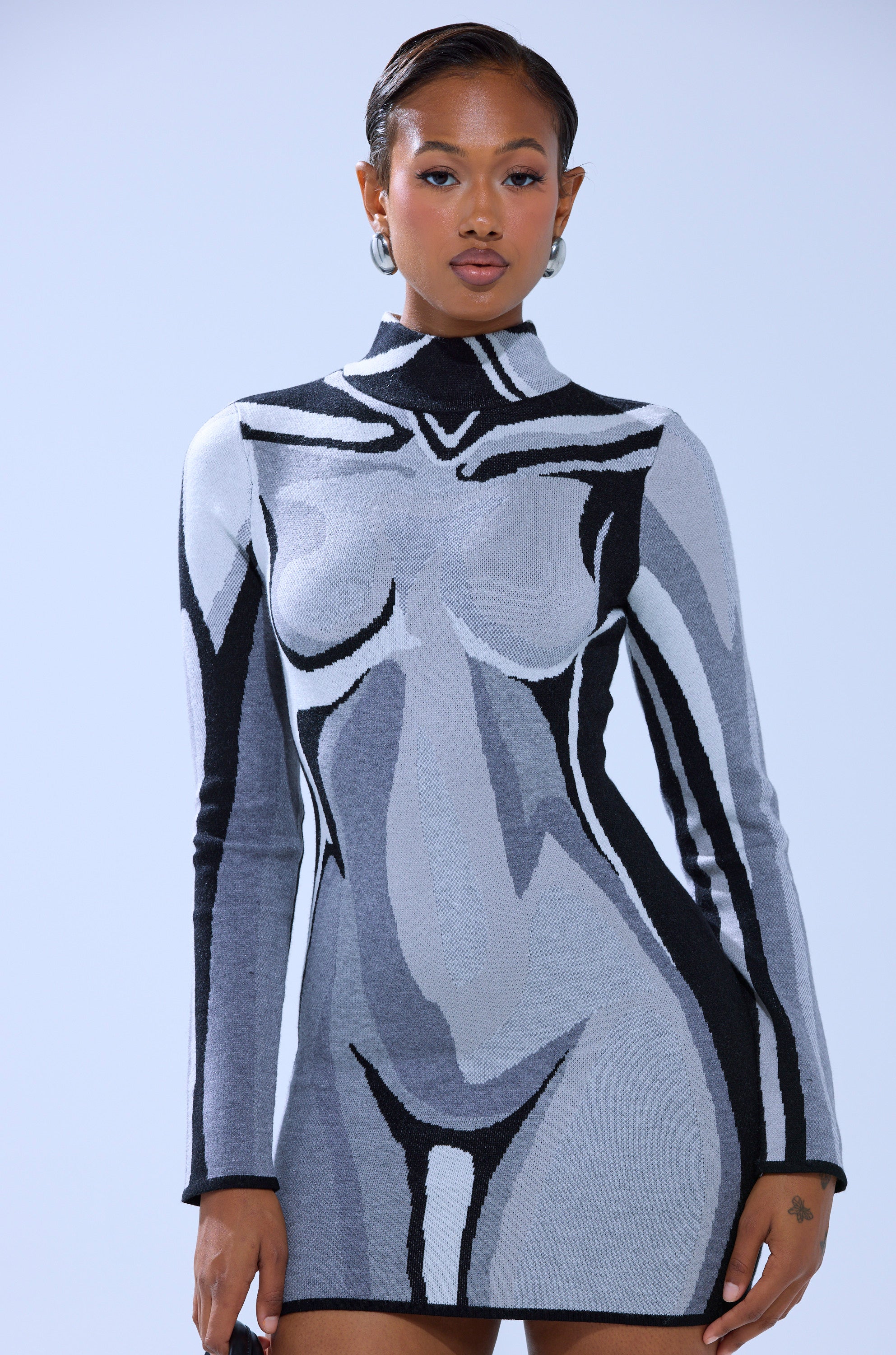 A woman with short hair and hoop earrings wears the A LIGHT SERVE PRINTED MINI DRESS, a curve-hugging knit featuring a bold gray and black abstract body graphic that resembles a stylized nude form. She has a calm expression.