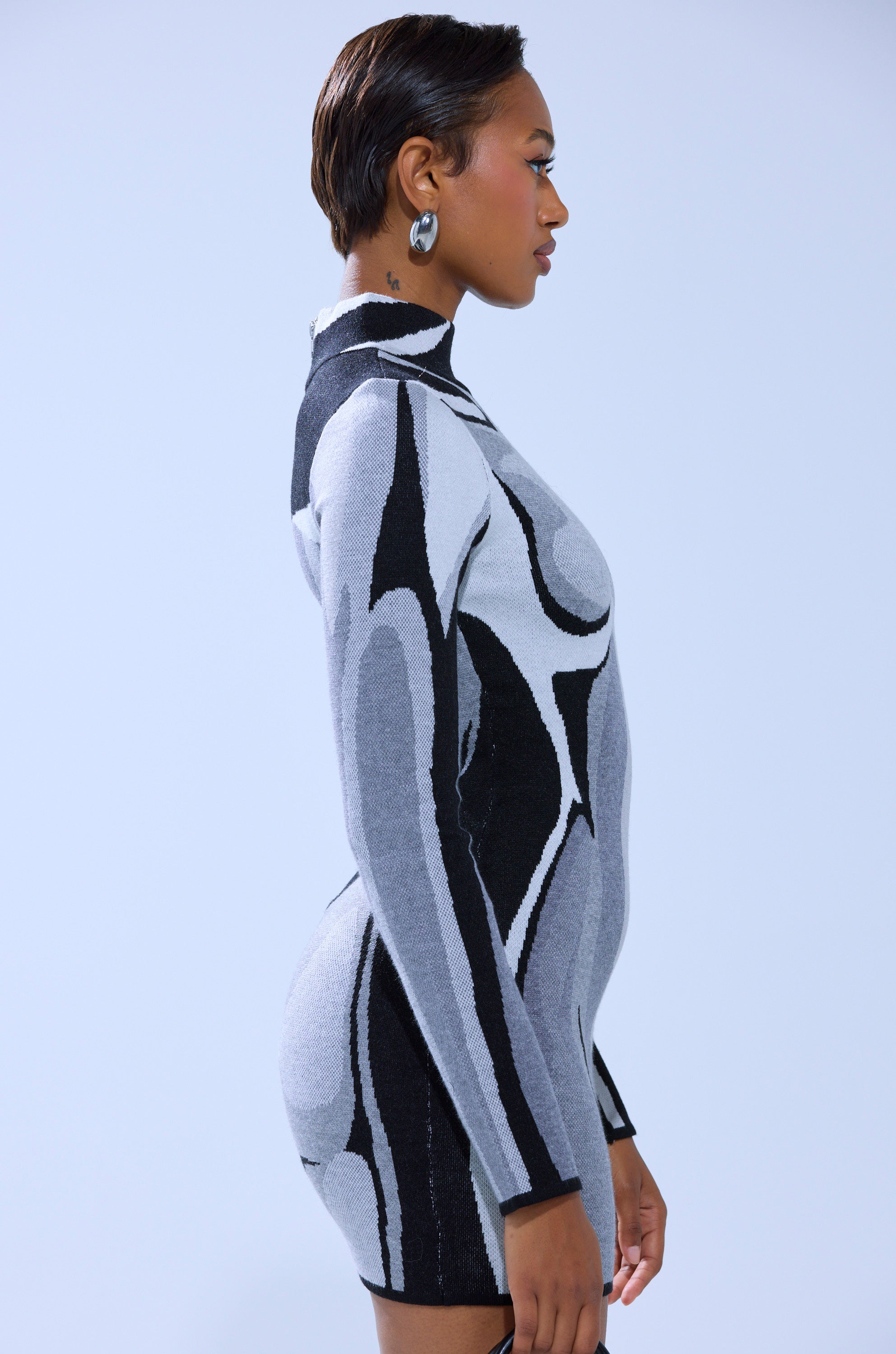 A person with short hair poses in profile, wearing the A LIGHT SERVE PRINTED MINI DRESS featuring an abstract black, white, and gray pattern, against a plain light blue background.