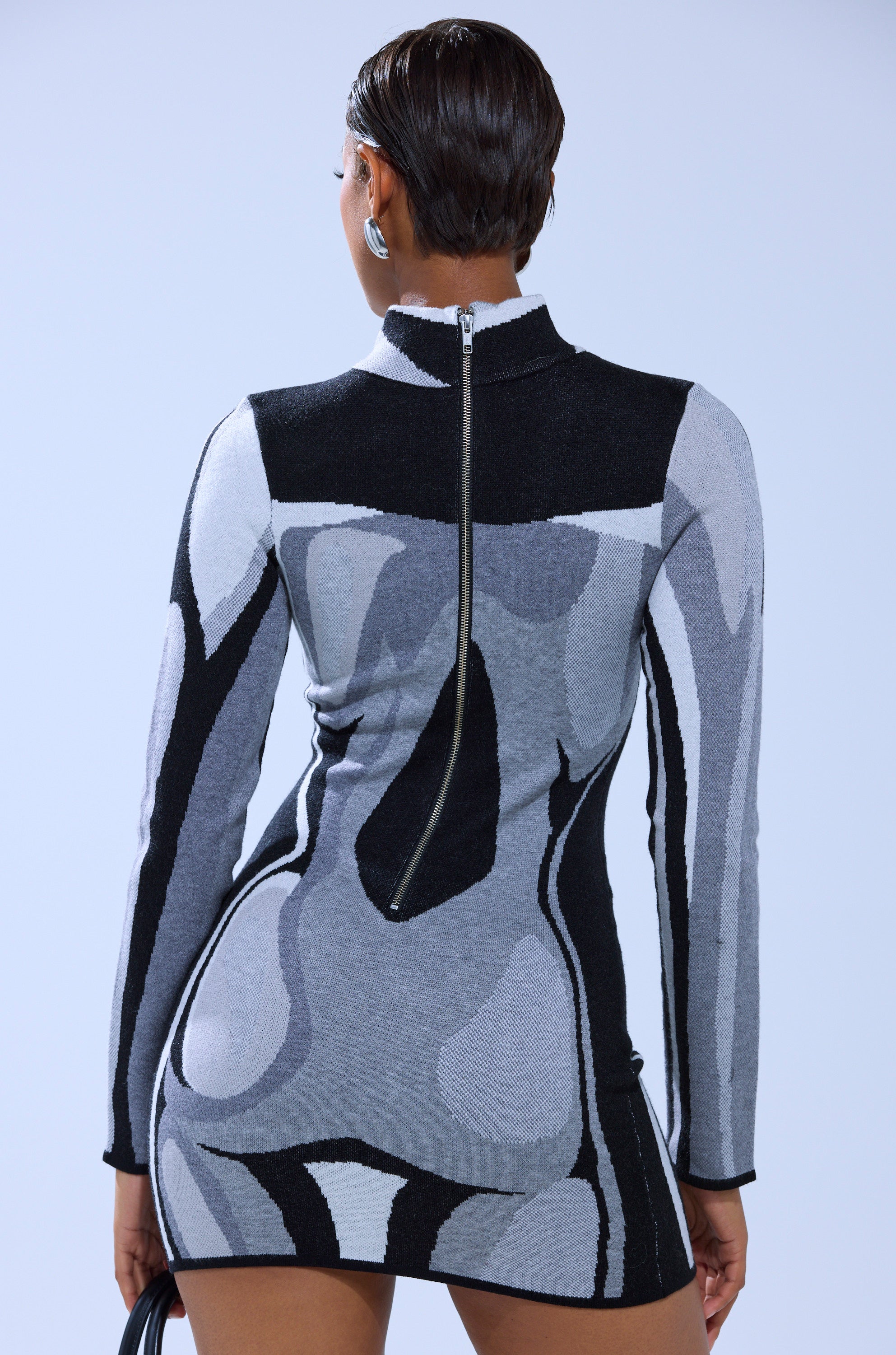 A person with short hair is shown from behind against a plain light background, wearing the A LIGHT SERVE PRINTED MINI DRESS featuring abstract gray, black, and white patterns and a visible back zipper.