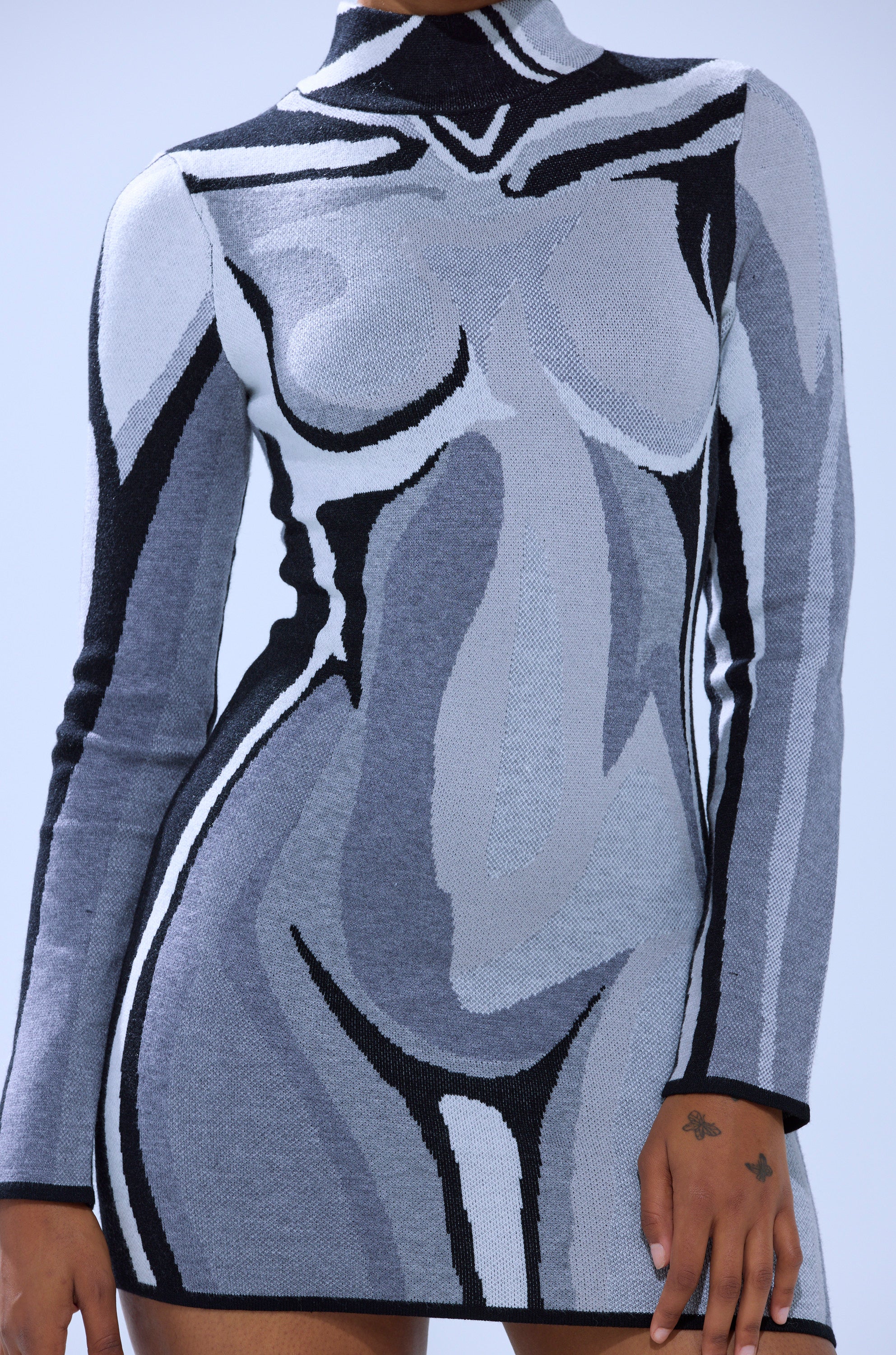 A person wears the A LIGHT SERVE PRINTED MINI DRESS, a form-fitting, long-sleeved mini dress featuring a monochrome abstract graphic outlining nude contours; one hand with small finger tattoos is visible.
