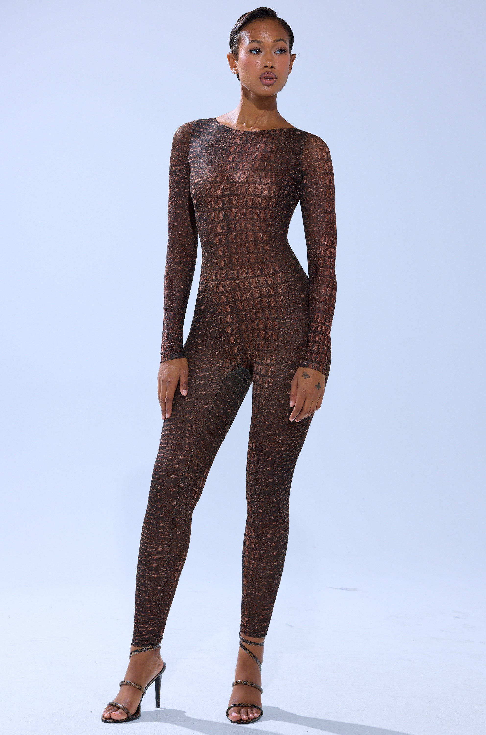 A woman stands confidently in the TRY AND GET ME SNAKE MESH JUMPSUIT—a fitted, long-sleeve brown snake print piece—paired with black high-heeled sandals against a plain light background.