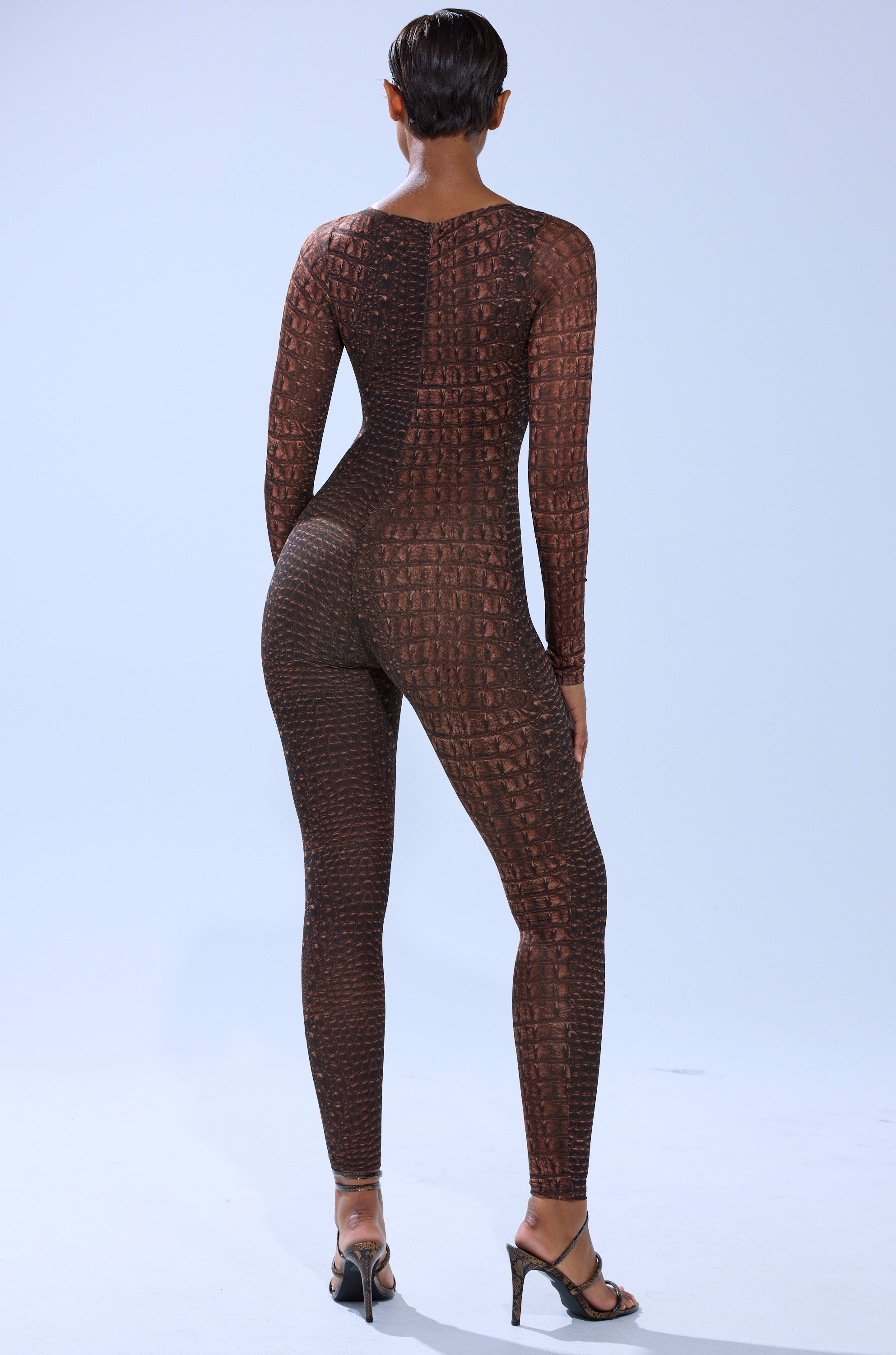 A person with short hair faces away against a plain background, wearing the fitted, long-sleeve brown TRY AND GET ME SNAKE MESH JUMPSUIT from AKIRA Label and high-heeled sandals.