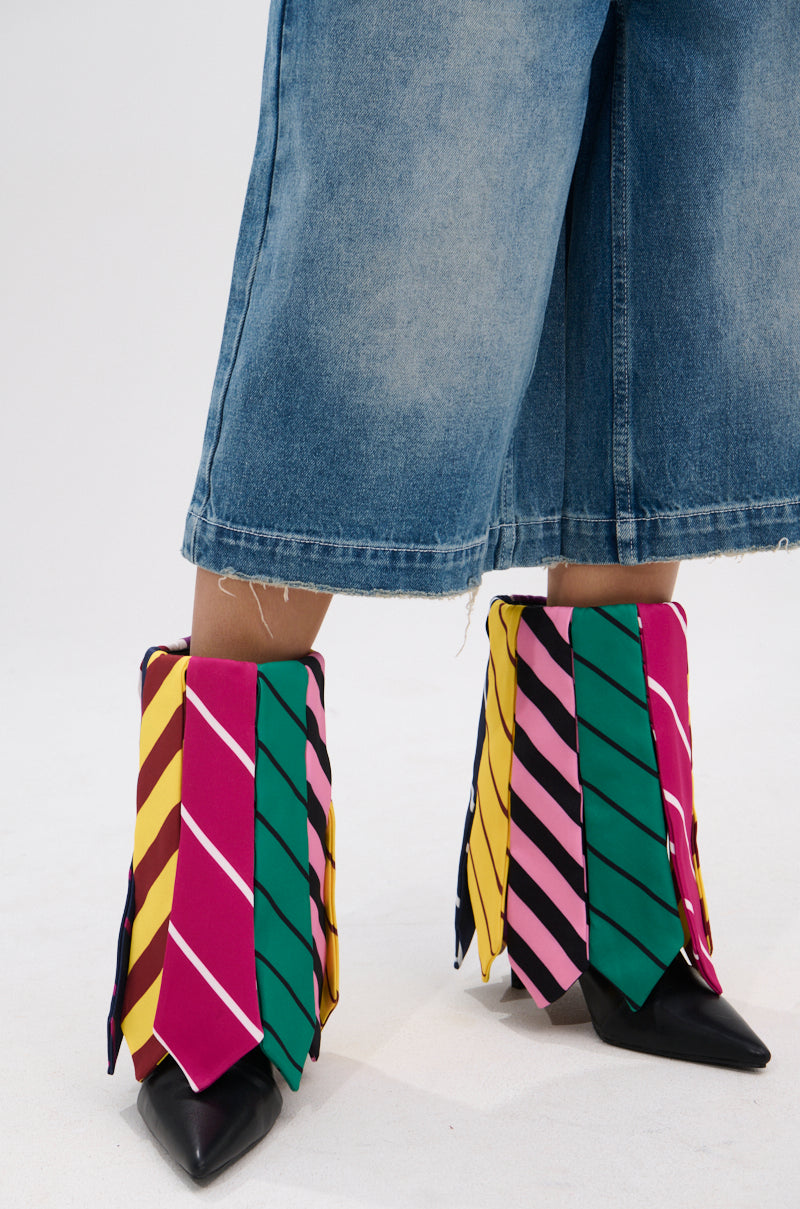 A person in wide, cropped denim jeans wears the AZALEA WANG JAUNE BLACK TIE BOOTIE—black, pointed-toe shoes uniquely adorned with colorful pink, green, and yellow neckties draped over them.