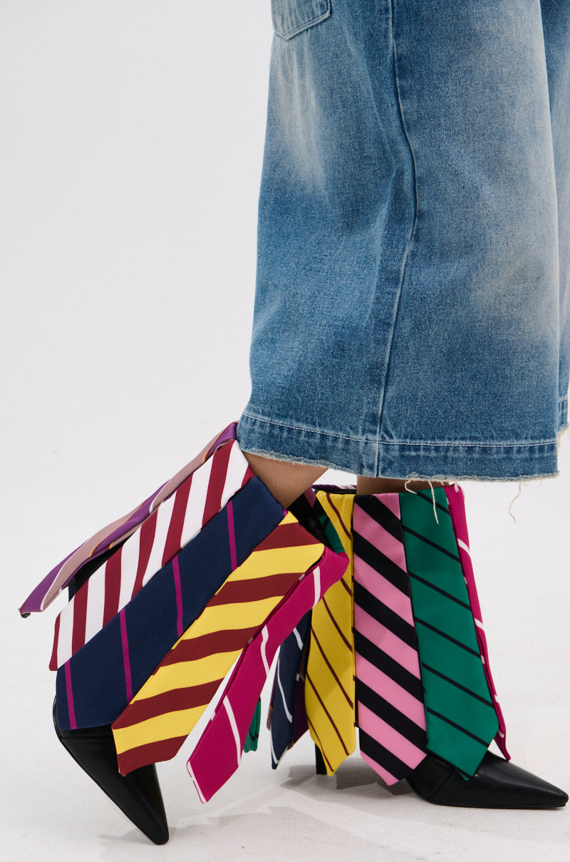 A person wears wide, cropped blue jeans and the AZALEA WANG JAUNE BLACK TIE BOOTIE, featuring several colorful, striped neckties draped over the chic ankle boots for a playful, unconventional fashion statement.