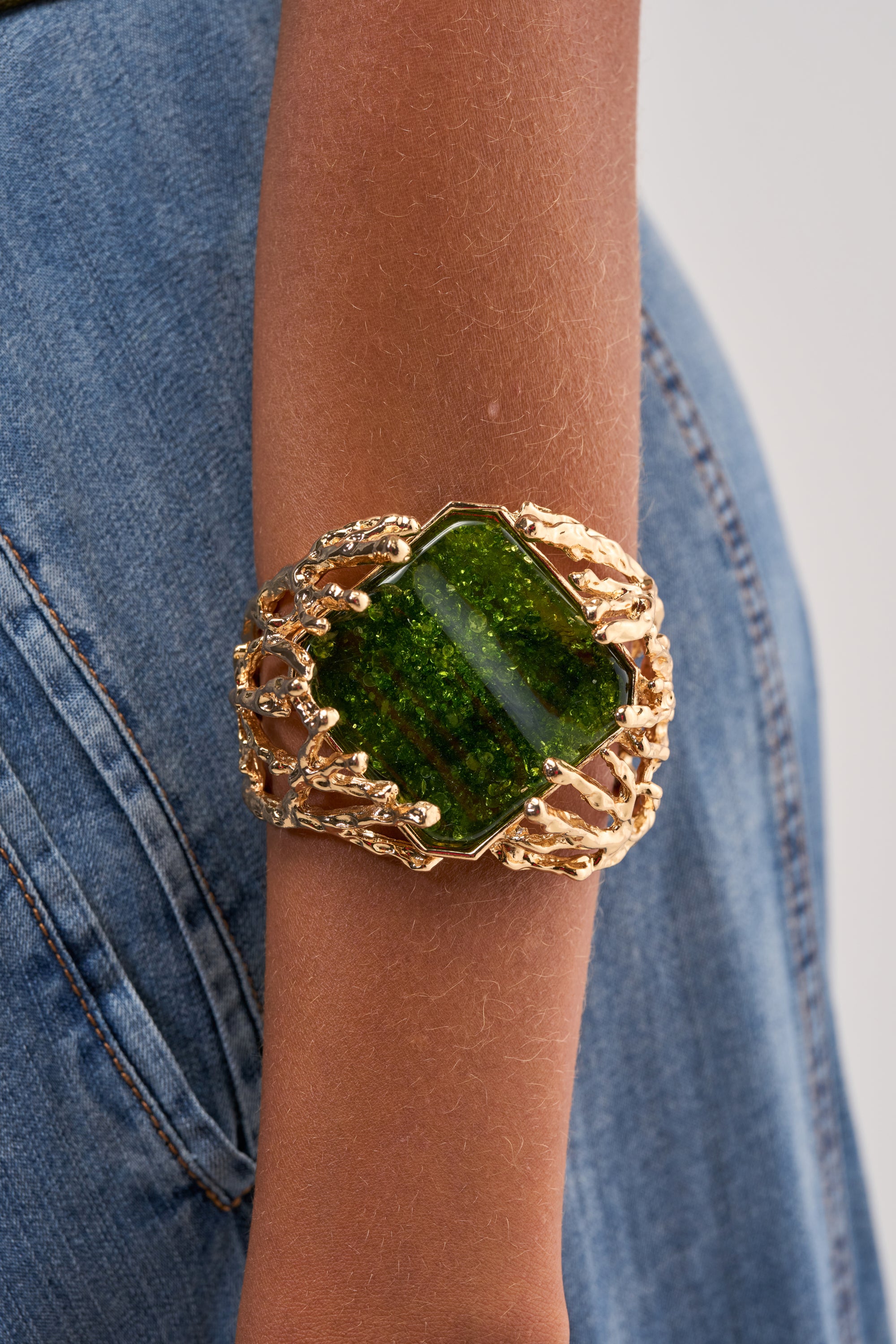 A person wearing the CHAMPAGNE DRIP GREEN GEM BRACELET, a large gold cuff with an ornate design and a central green gemstone, perfect as wedding guest jewelry, paired with blue denim clothing.