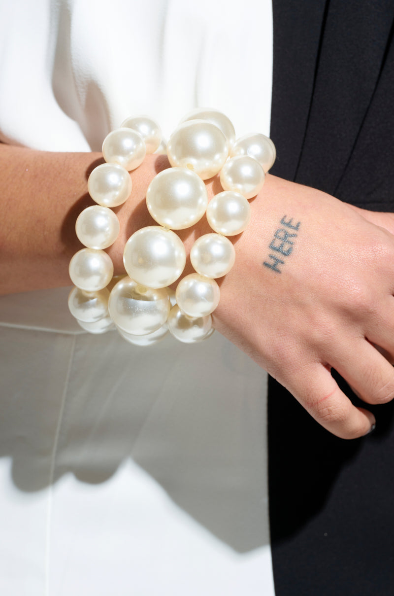 A person adorned with a PEARL PLEASE STRETCH BRACELET SET on their wrist, which features a polished pearl-like design. The word "HERE" is tattooed along the side of their hand. Their attire, composed of black and white fabric, highlights a sleek yet stretchy aesthetic. 1
