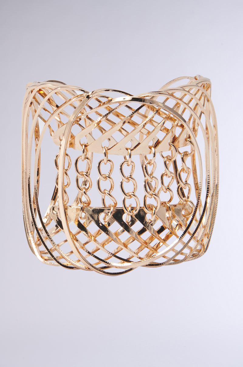 The CHAIN CHIC BRACELET features a faux gold metallic finish with intricately crisscrossed metal bands and detailed chain link accents, offering an elegant and textured appearance against a simple backdrop.